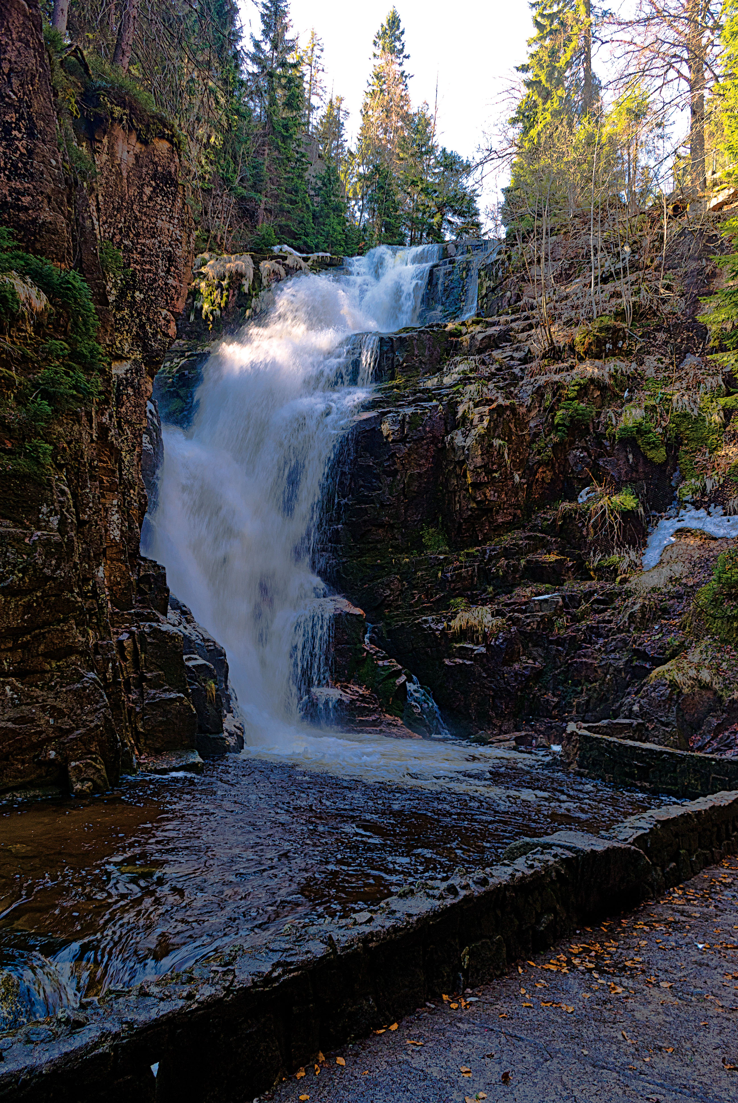 Waterfall flowing over rocky cliffs surrounded by forest with stone pathway in foreground
