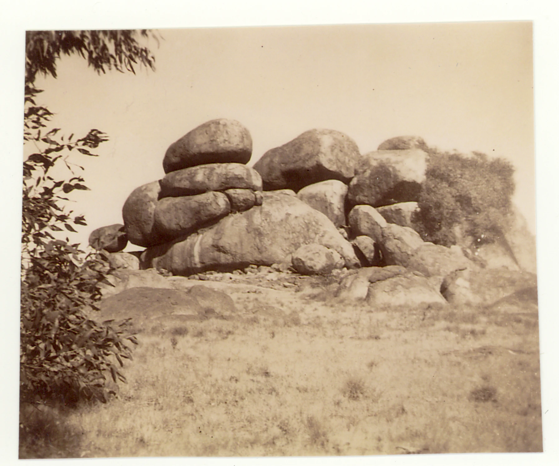 A sepia-toned vintage photograph showing a cluster of large, weathered, rounded granite boulders in an arid landscape with a small shrub in the foreground left.