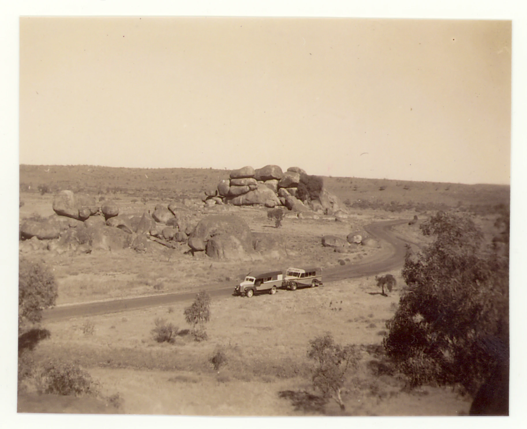A sepia-toned vintage photograph showing a vehicle driving along a road past large, stacked granite boulders in an arid Australian landscape.