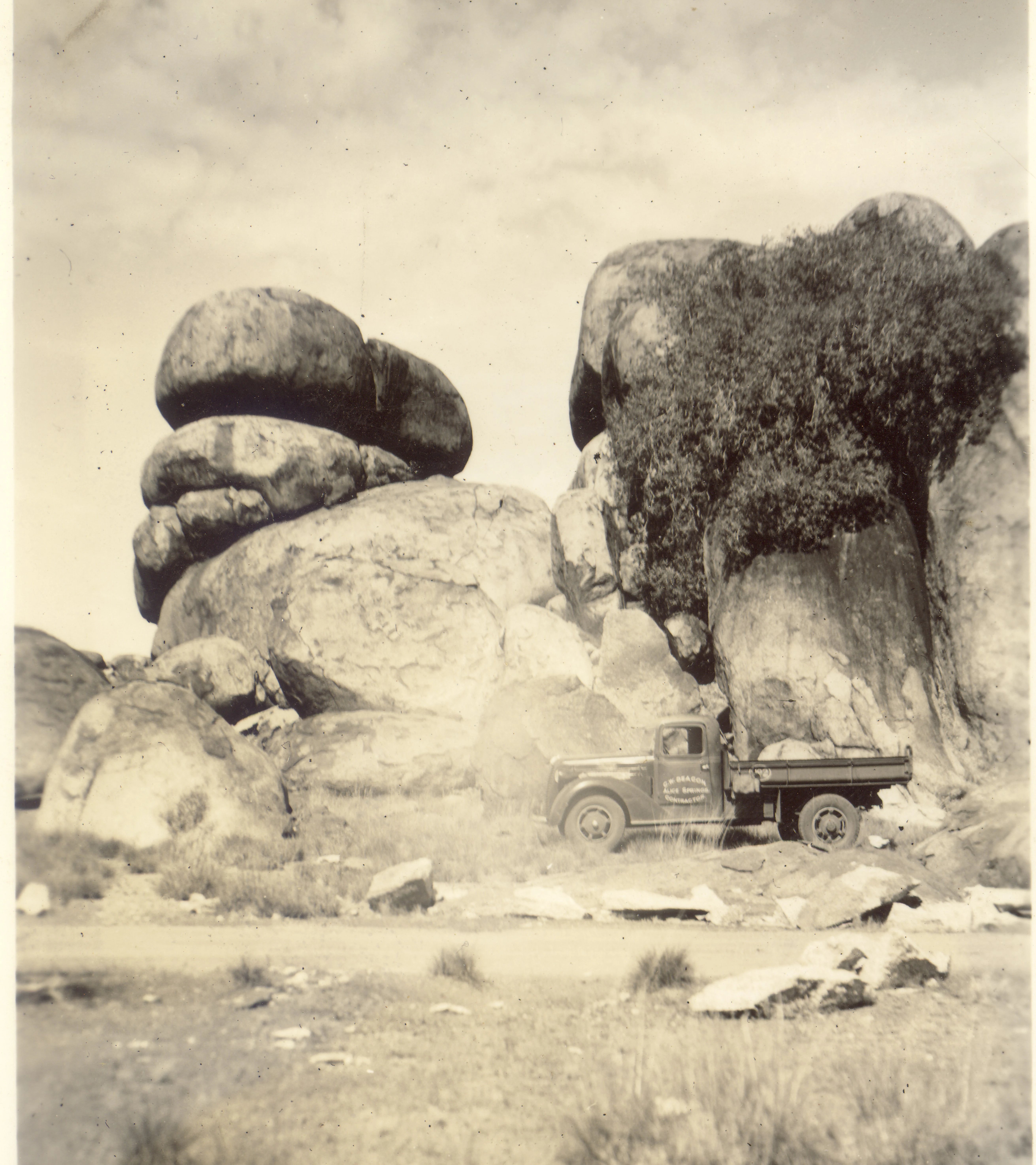 A black and white vintage photograph showing a truck parked in front of large, rounded granite boulder formations under a cloudy sky.
