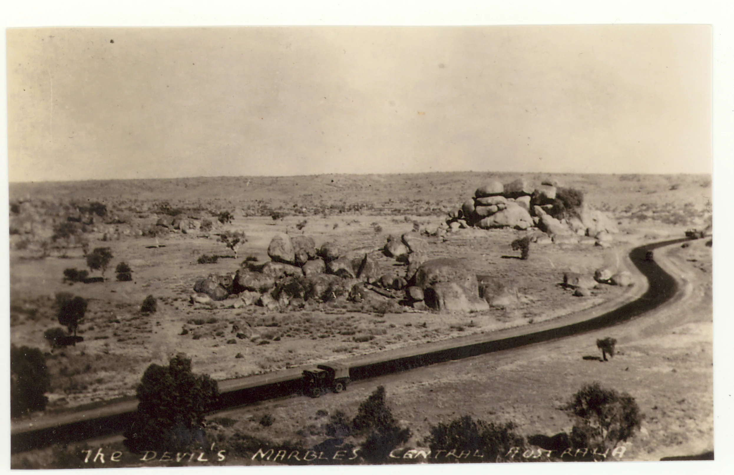 A black and white historical photograph shows large rounded granite boulders scattered across a dry, scrubby landscape, with a road curving through the foreground and text at the bottom reading The Devil's Marbles, Central Australia.