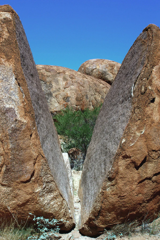 A close-up perspective of two massive granite boulders leaning against each other, creating a V-shaped gap that reveals green shrubbery and a clear blue sky in the background.
