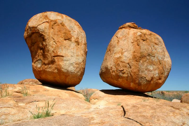 Two large rounded orange granite boulders balanced on top of a rocky outcrop against a clear blue sky.