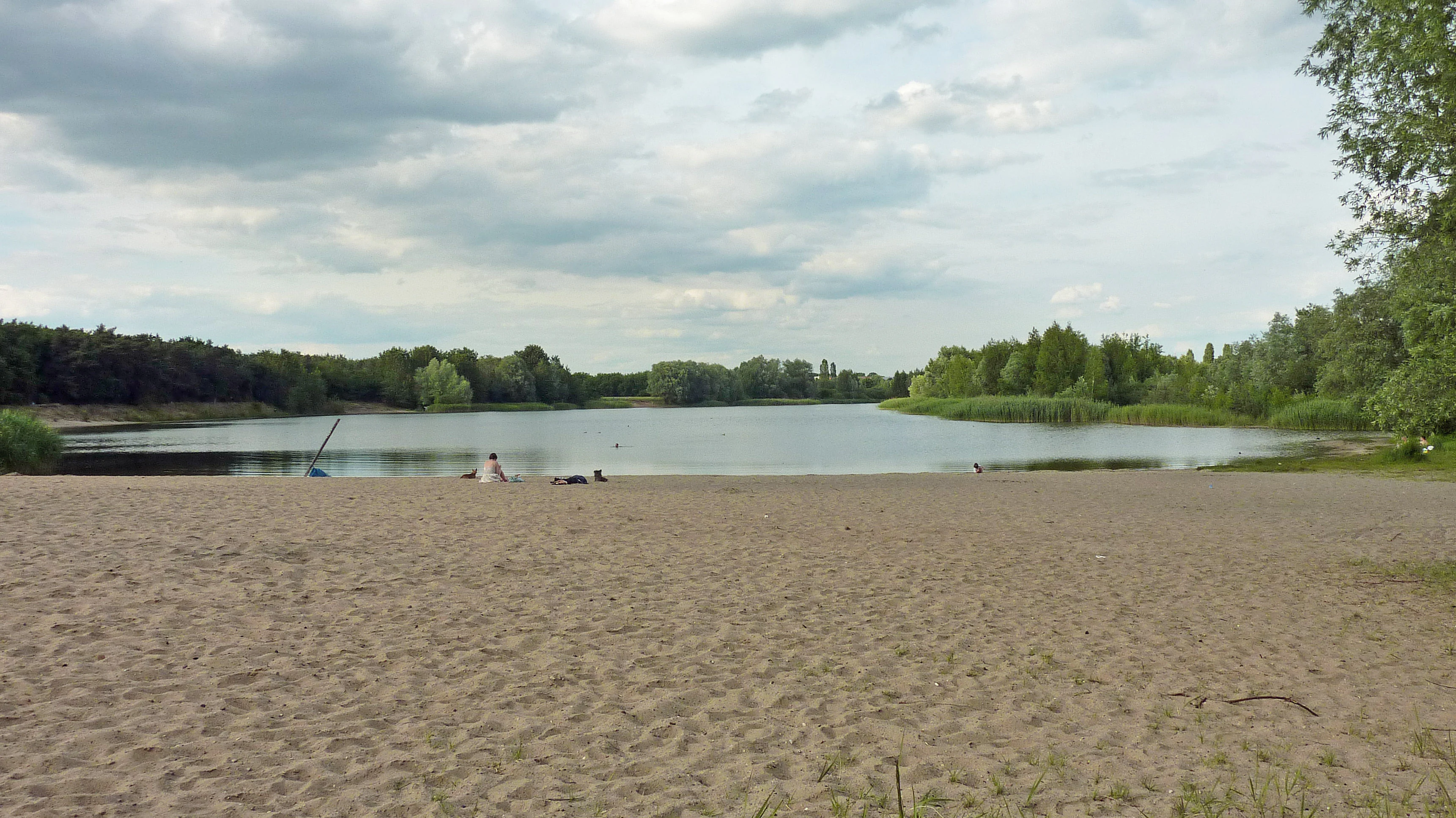 A wide shot of a sandy beach in the foreground leading to a calm lake, with forested shorelines and a cloudy sky in the background.