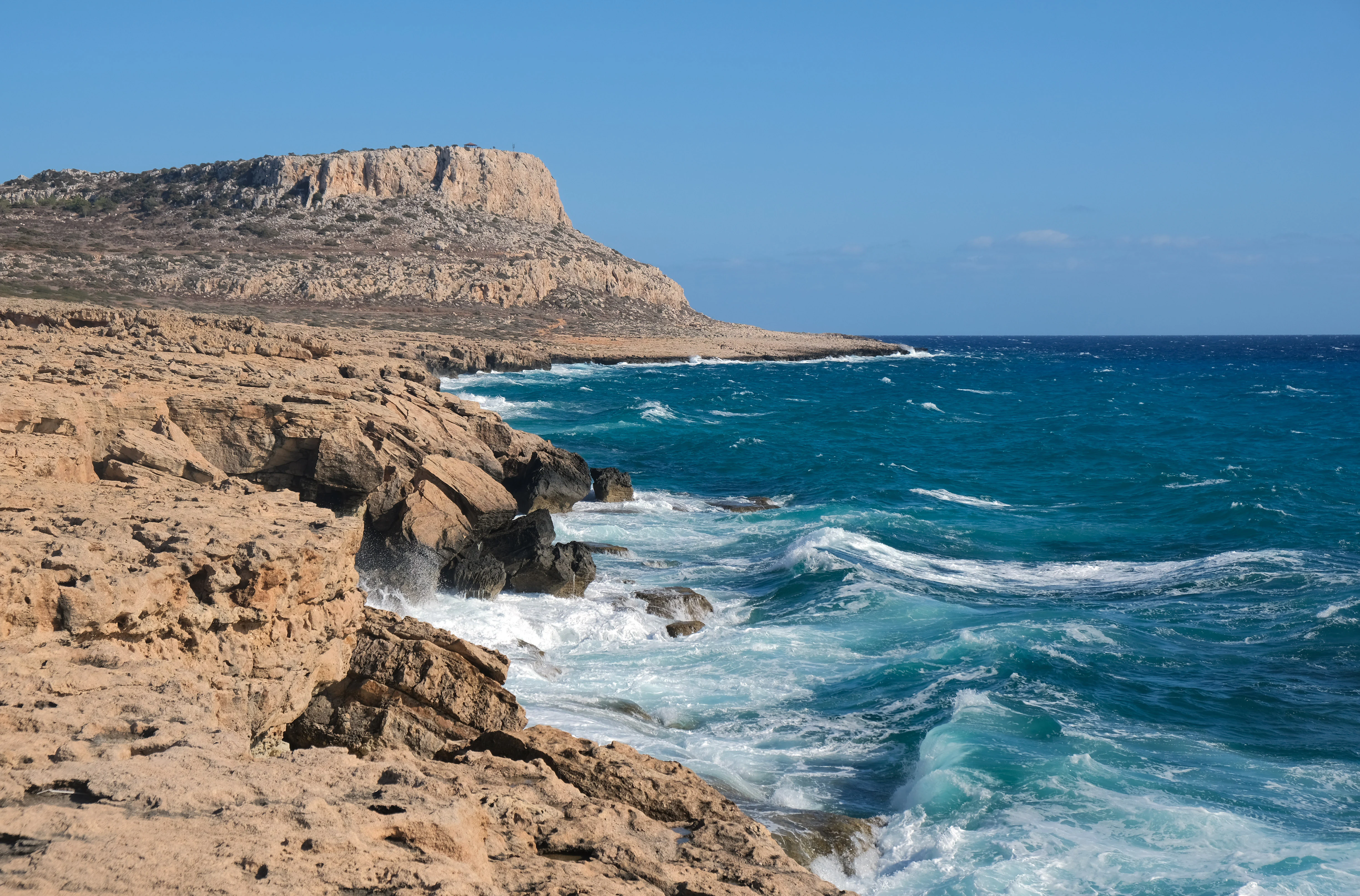 A wide coastal landscape showing jagged, light brown rocky cliffs meeting the deep blue Mediterranean Sea with white waves crashing against the base.