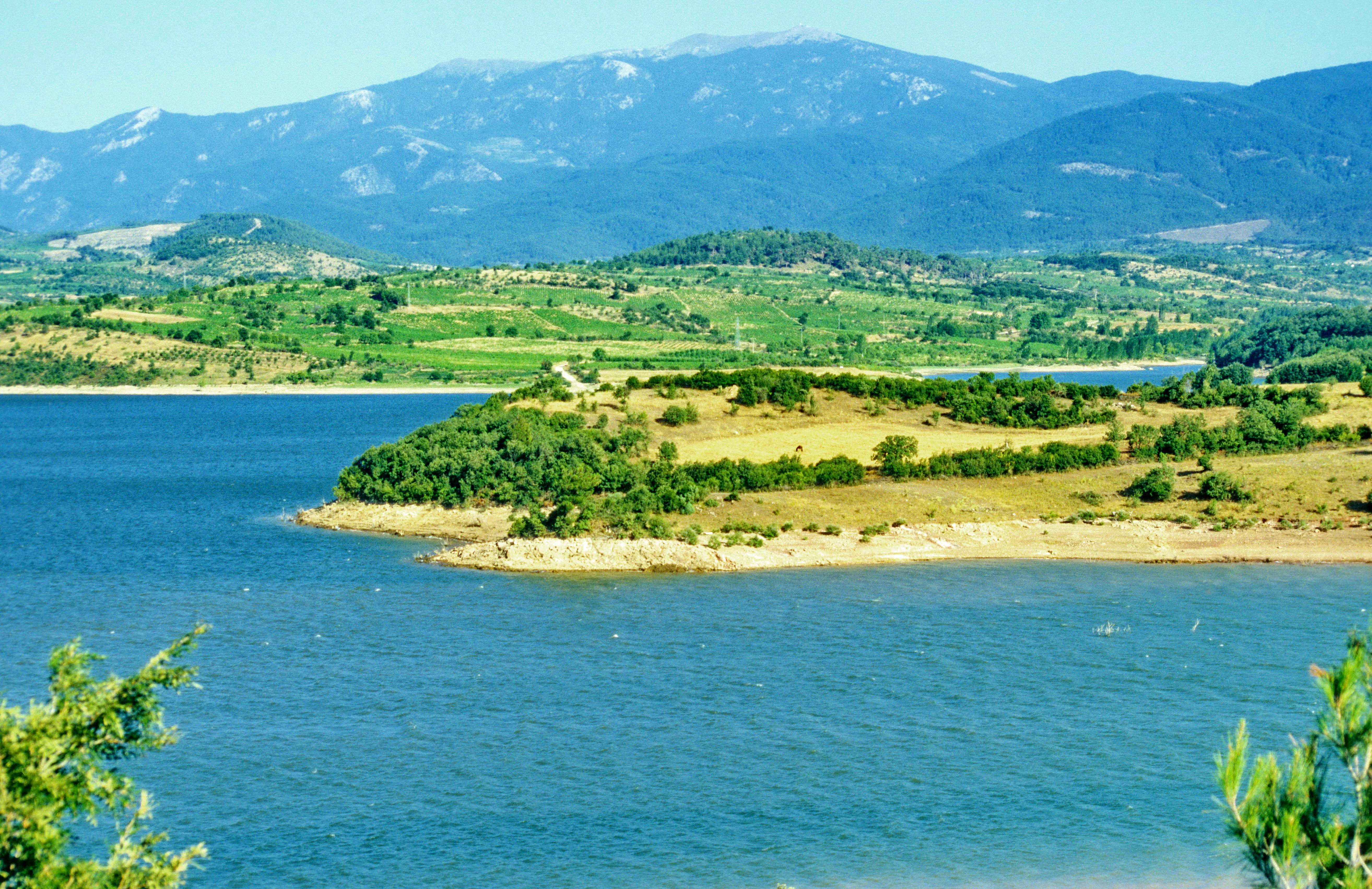 Wide view of a large reservoir with shoreline, green fields, and mountains in the background