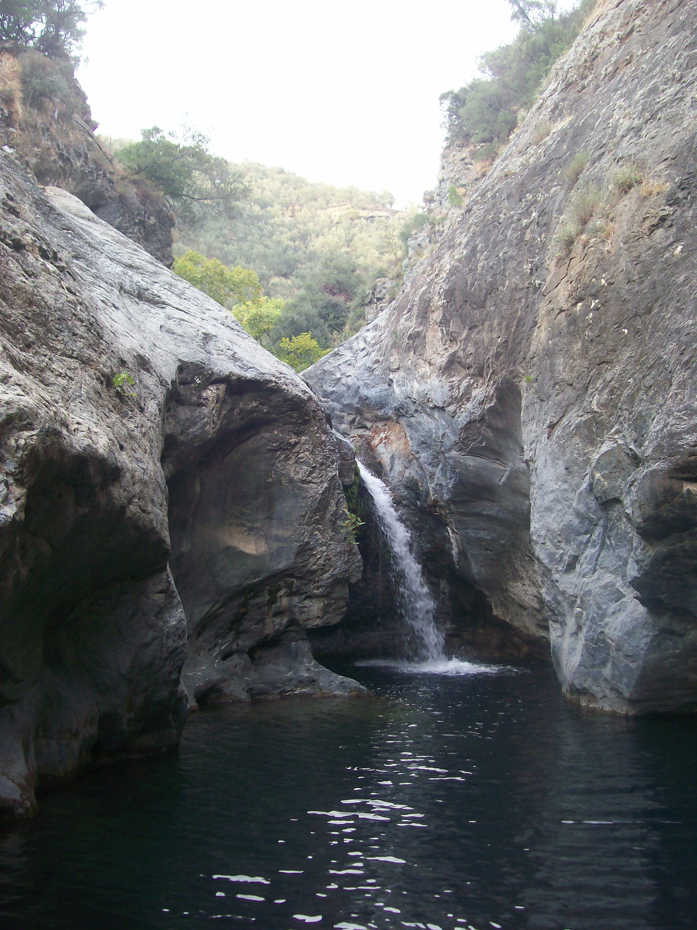 Waterfall flowing between large rocky cliffs into a dark pool of water with green vegetation in the background