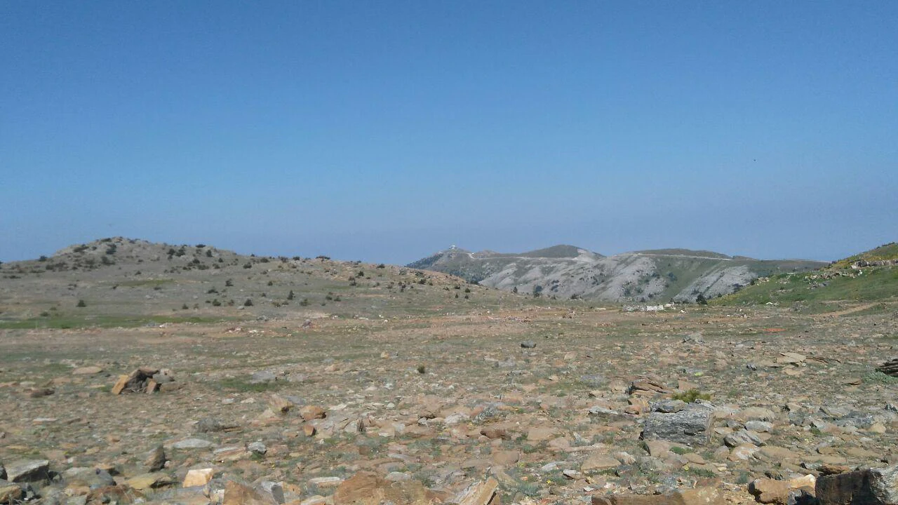 Wide rocky plateau with scattered stones and sparse vegetation, distant mountains under clear blue sky