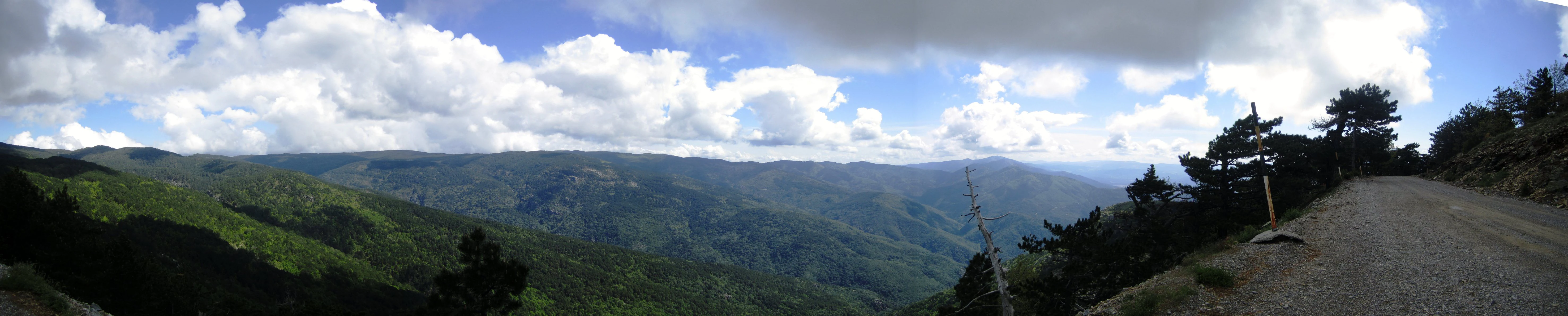 Panoramic landscape of mountainous terrain with rolling valleys, a gravel road, scattered trees, and a signpost under a partly cloudy sky