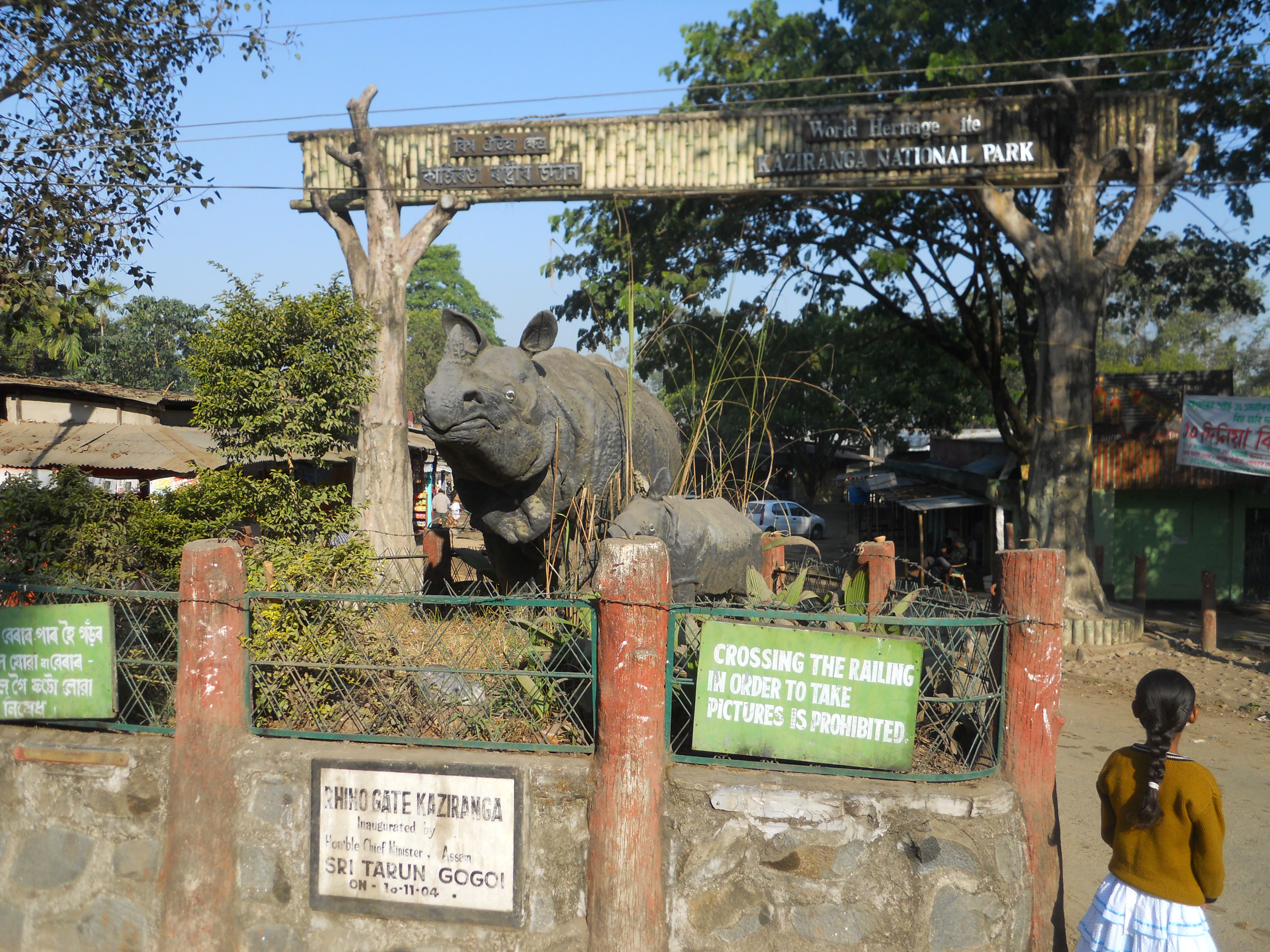 Entrada de muro de piedra con pilares rojos, letreros verdes y una gran estatua de rinoceronte en el Parque Nacional de Kaziranga. Una persona está cerca de la entrada.