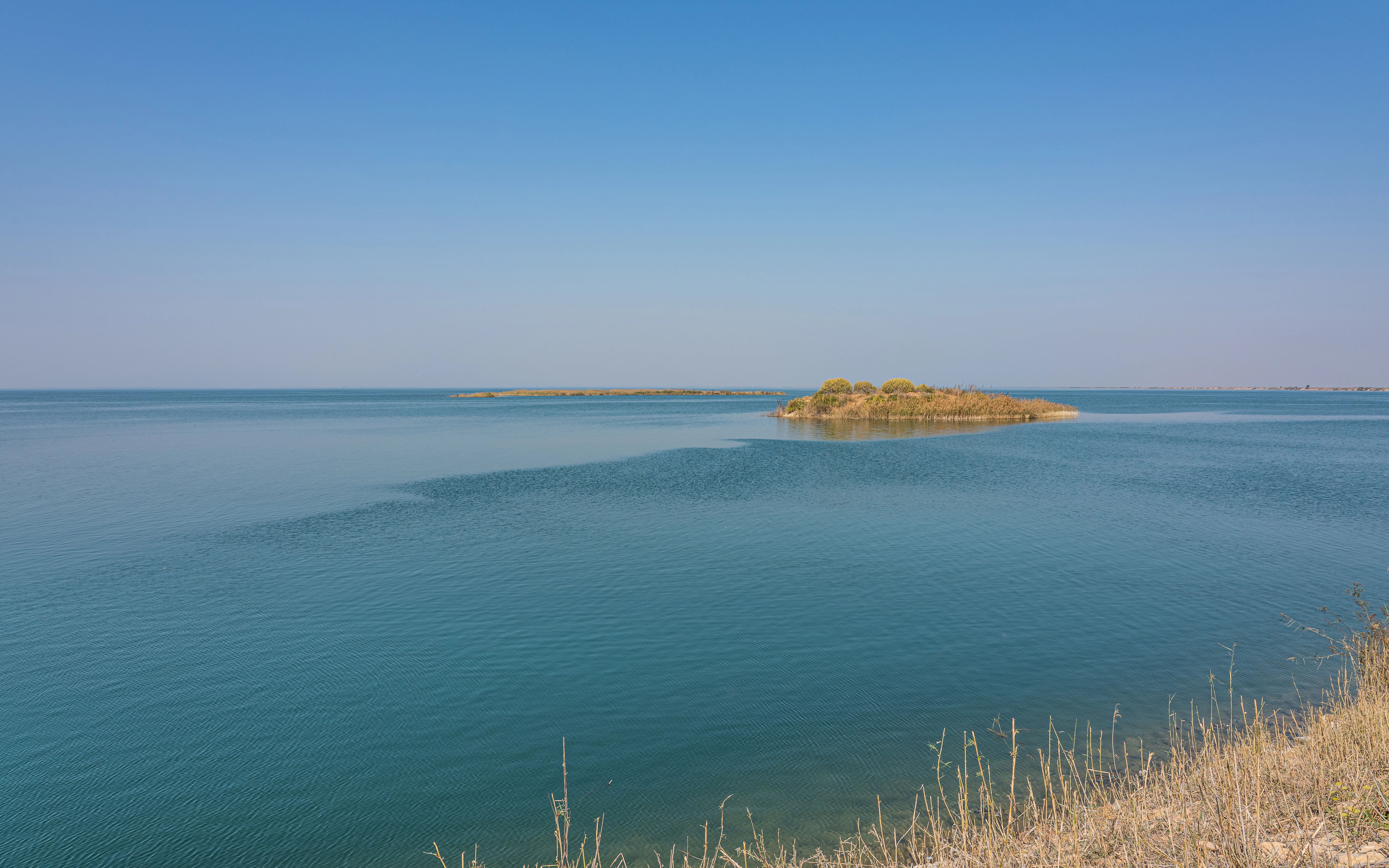 A wide, calm, blue lake with small, low-lying islands in the distance under a clear, bright blue sky.