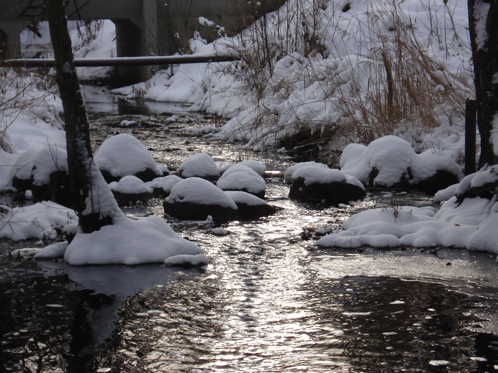 Snow-covered river with rocks and trees, bridge in background, winter scene