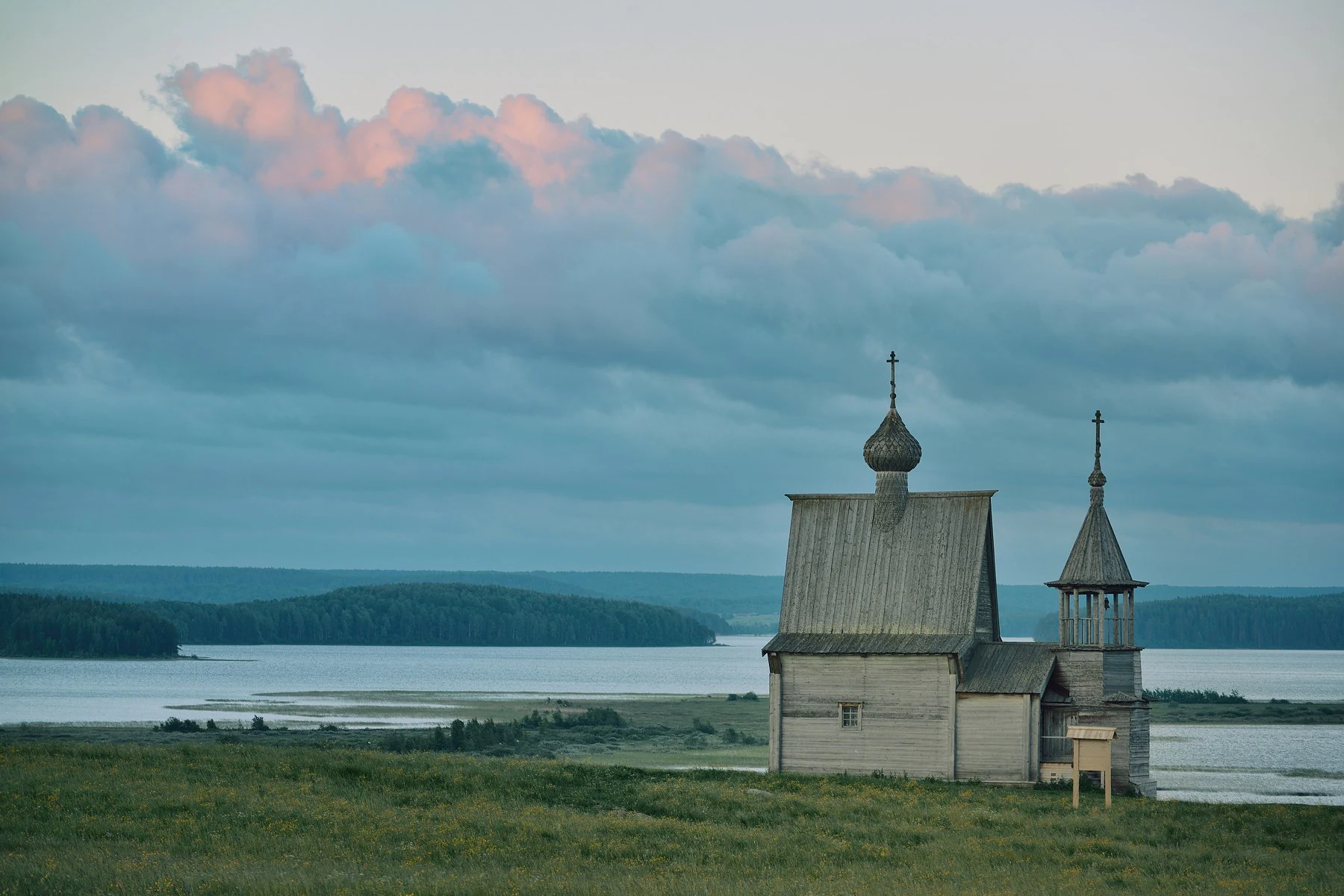 Wooden church with onion domes standing on a grassy field by a lake under a cloudy sky with pinkish clouds