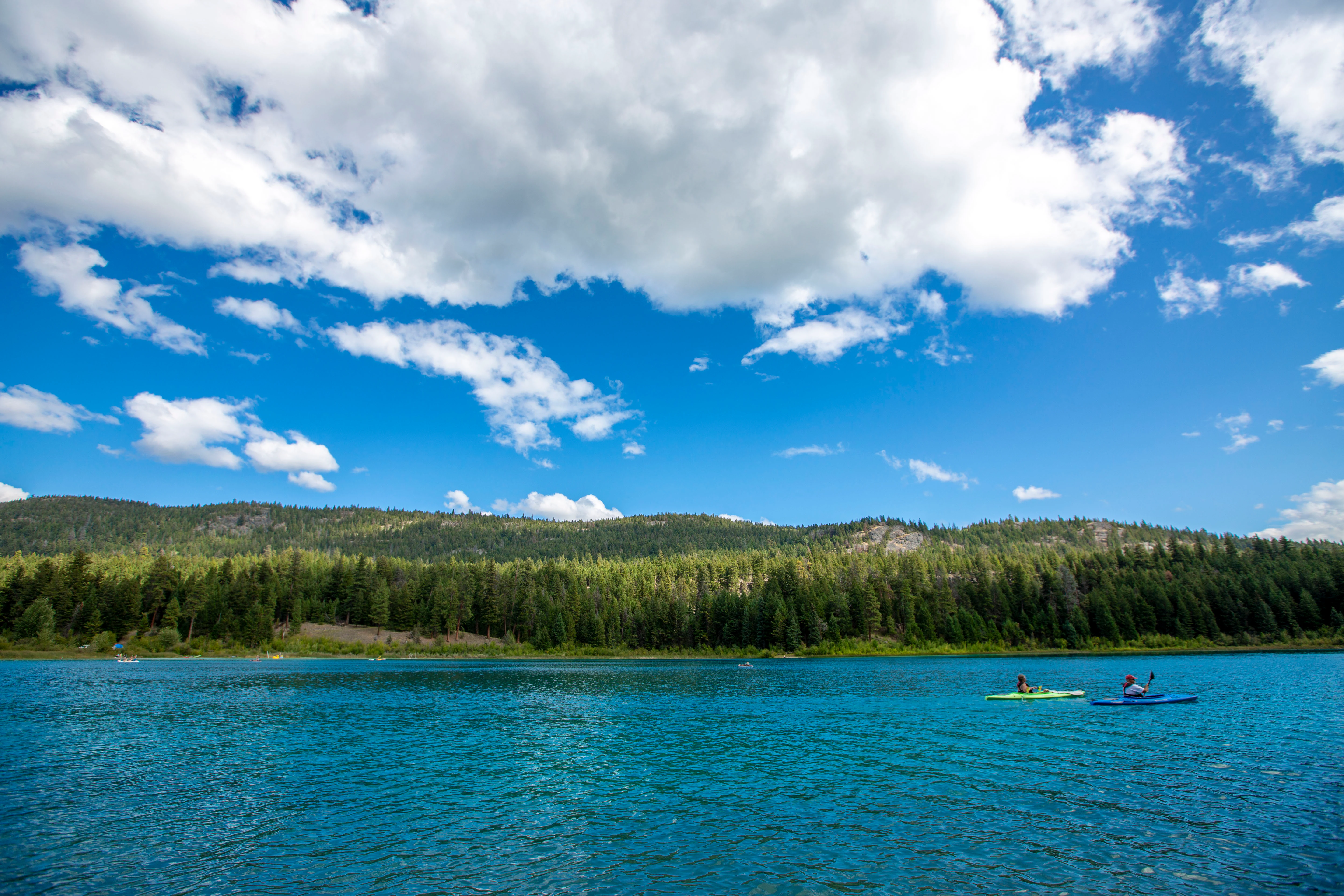 A wide, eye-level shot of a bright turquoise lake with two kayakers in the distance, backed by a dense green pine forest and rolling hills under a blue sky with fluffy clouds.