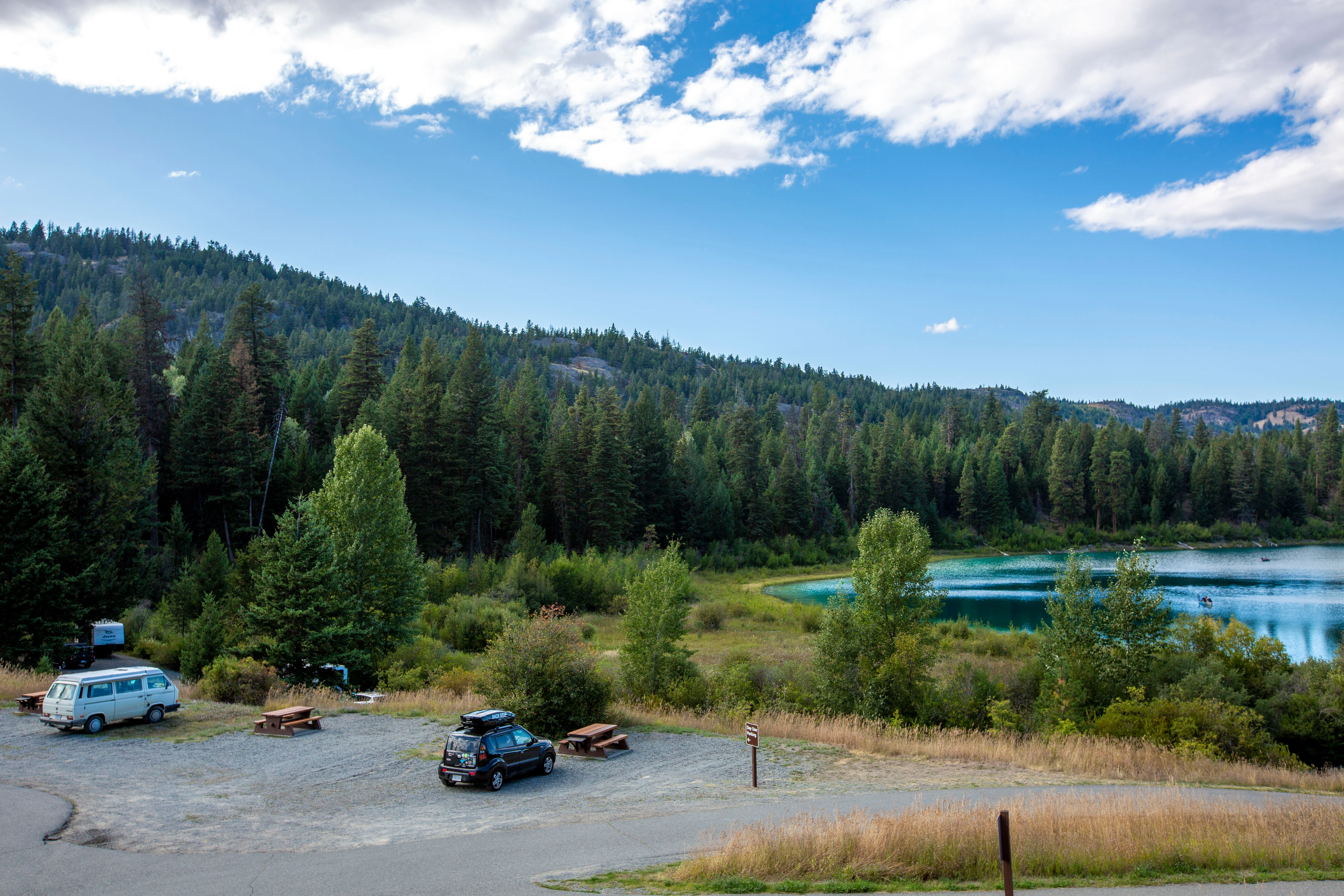 A gravel parking area with two vehicles and wooden picnic tables, bordered by a dense conifer forest and a turquoise lake under a bright blue sky with white clouds.