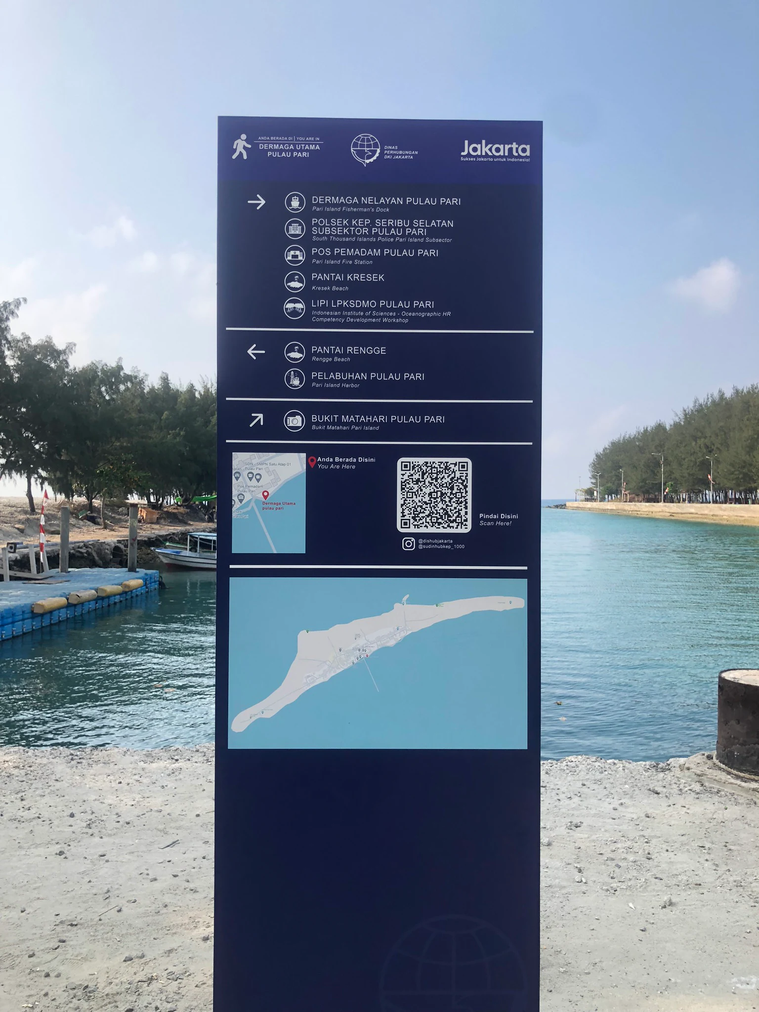 Tall dark blue signboard with directional text, QR code, and map of Pulau Pari, set against a coastal backdrop with water, trees, and boats