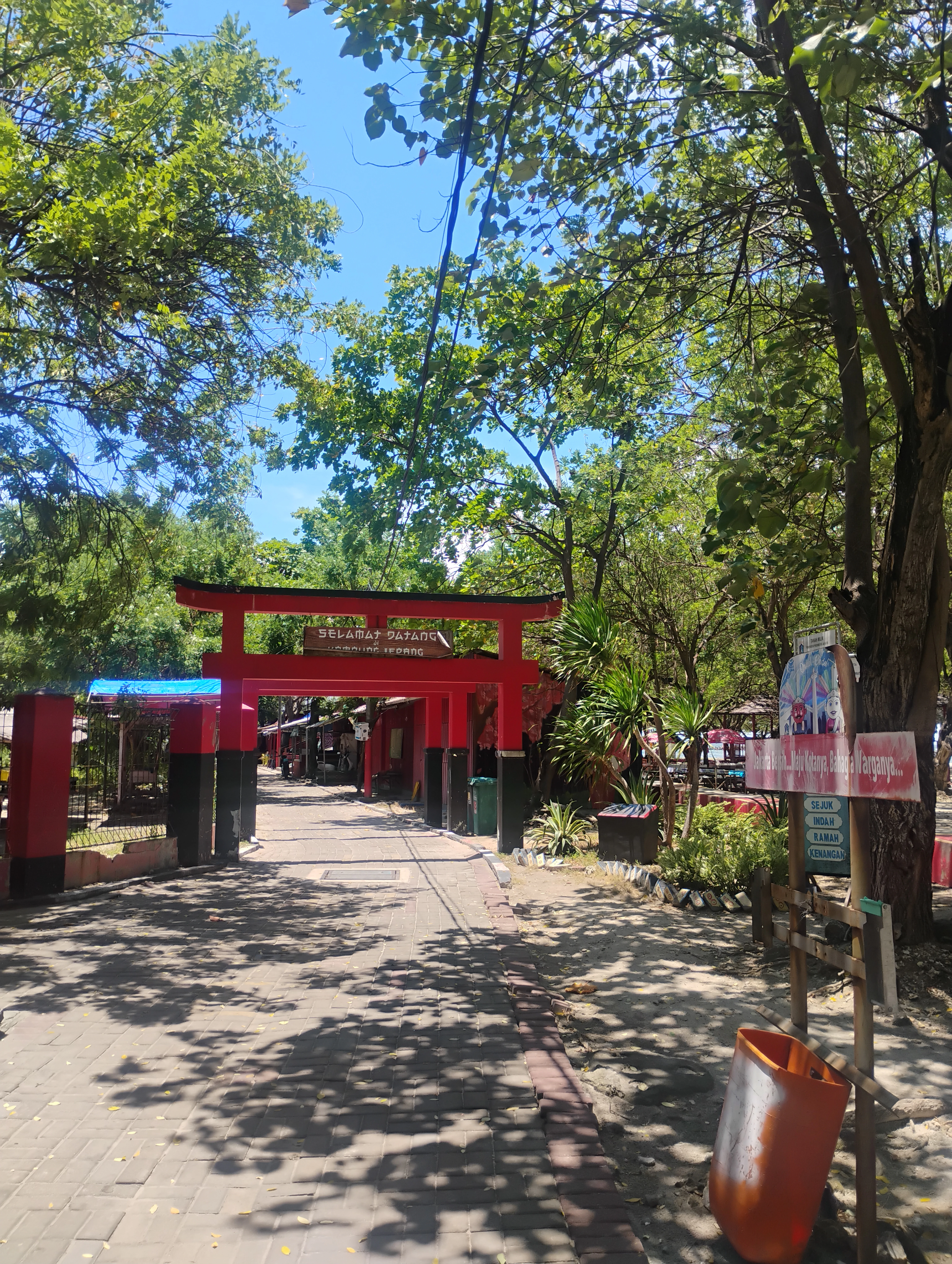 Red traditional gate structure at park entrance with paved pathway, trees, and signboard under clear blue sky