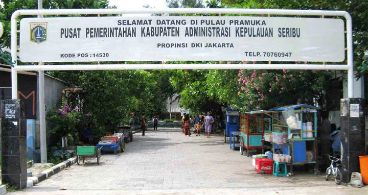 Large white sign reading 'SELAMAT DATANG DI PULAU PRAMUKA' and 'PUSAT PEMERINTAHAN KABUPATEN ADMINISTRASI KEPULAUAN SERIBU' with people walking on a paved path, trees, and a food cart in the background