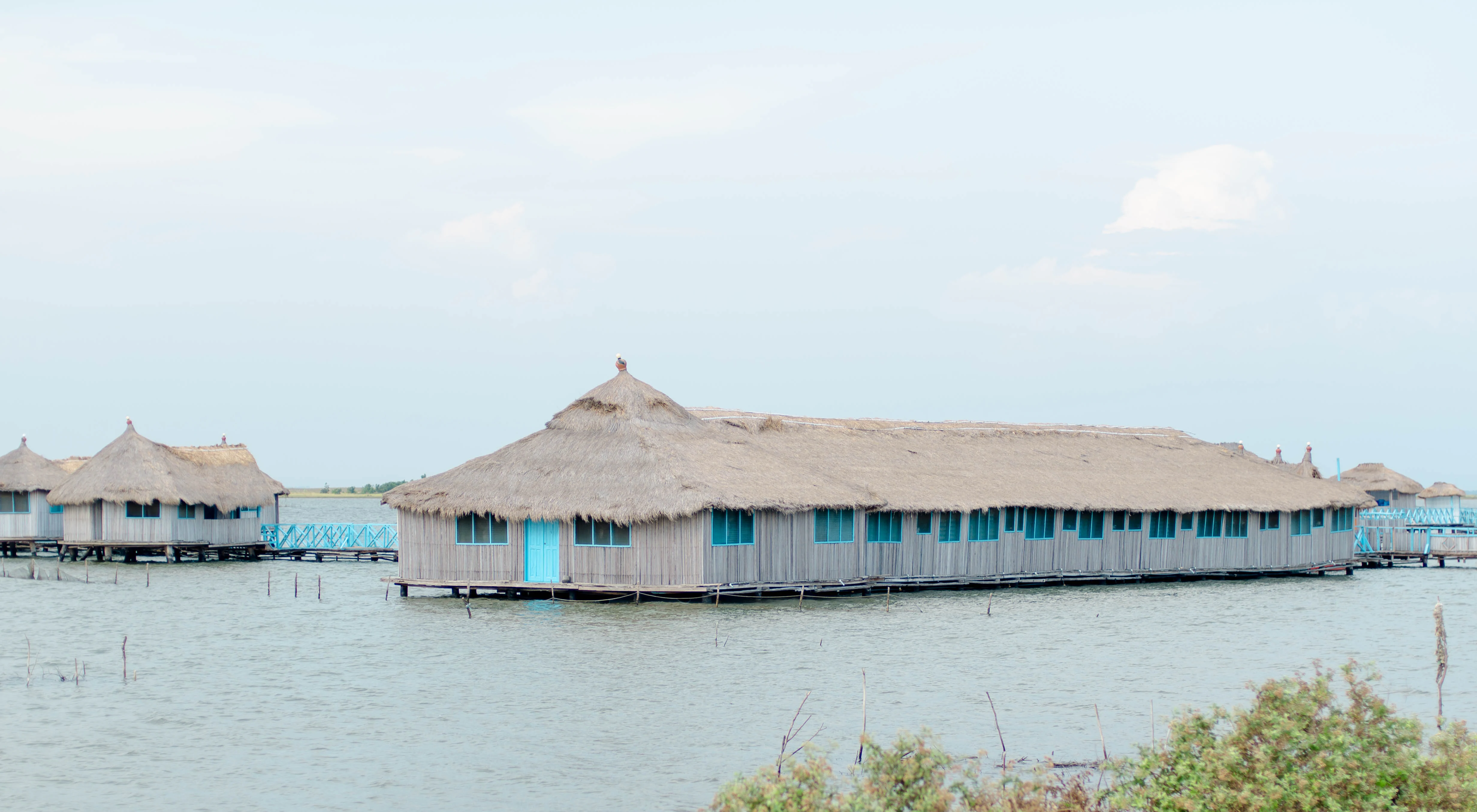 A series of wooden buildings with thatched roofs built on stilts over calm lagoon water under a pale sky.