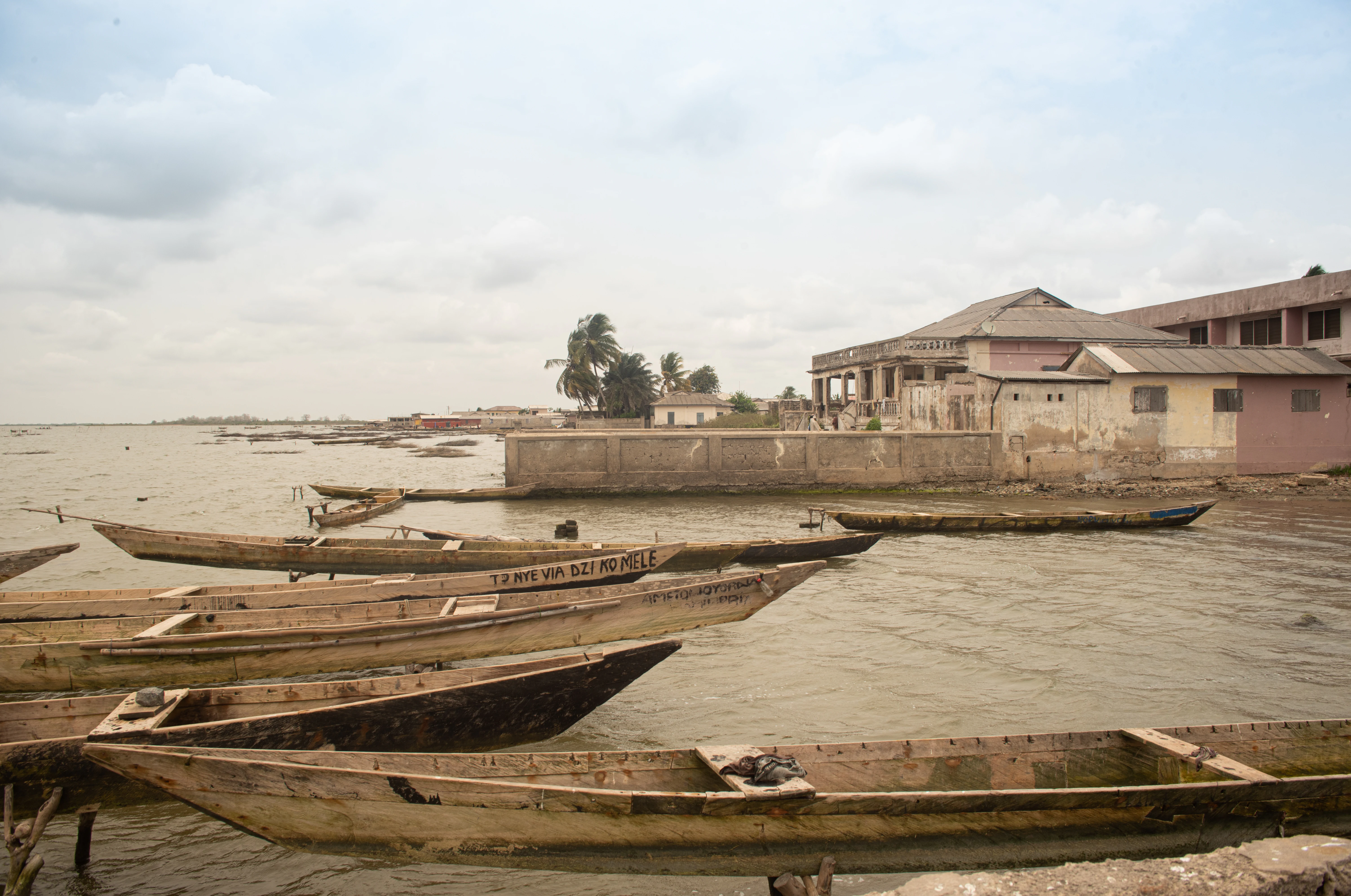 A group of weathered wooden canoes docked in shallow water near buildings on the shore of Keta Lagoon.