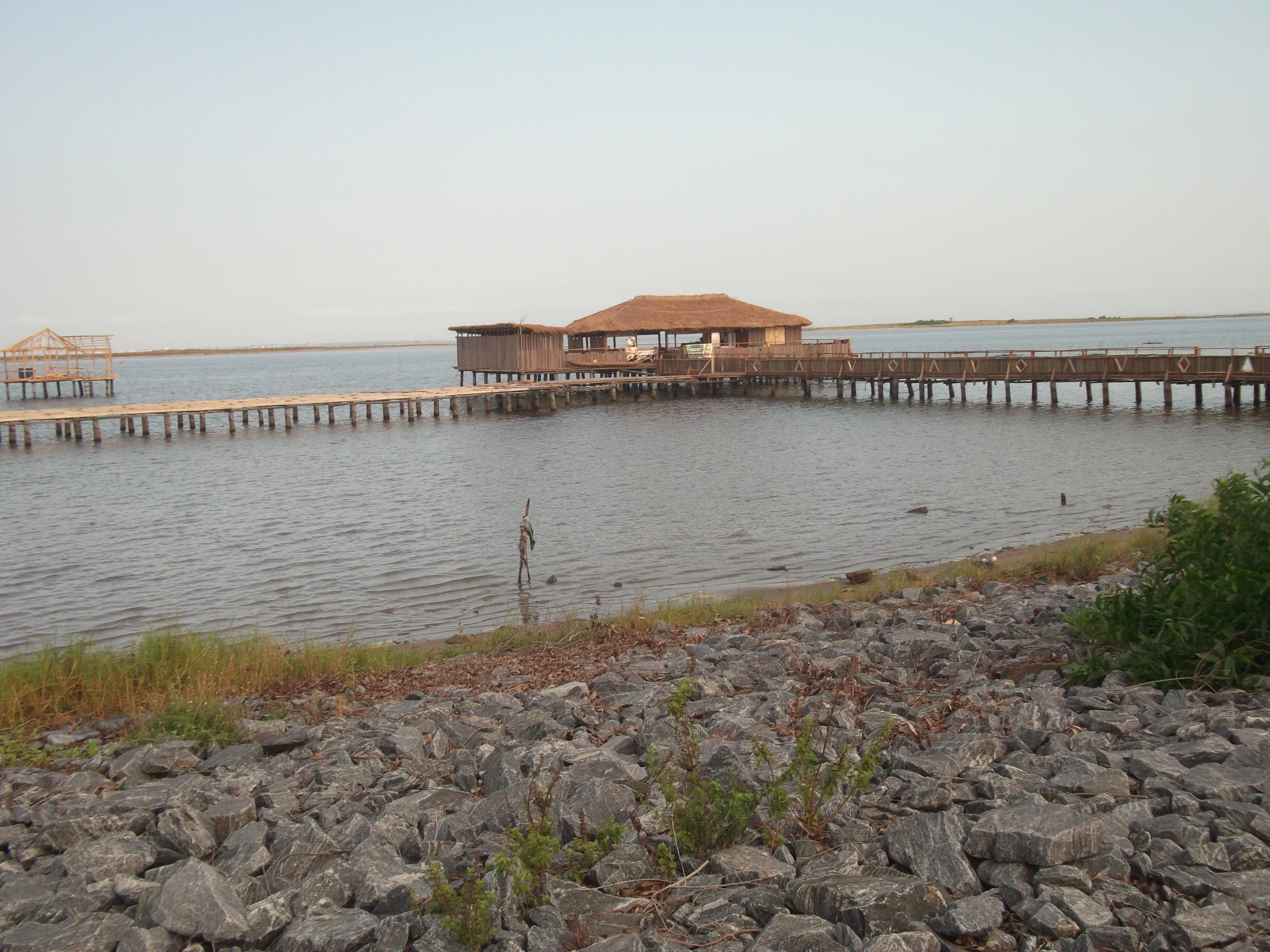 A long wooden pier with a roofed structure at its end stretches out over the calm water of Keta Lagoon, with a rocky shoreline in the foreground.