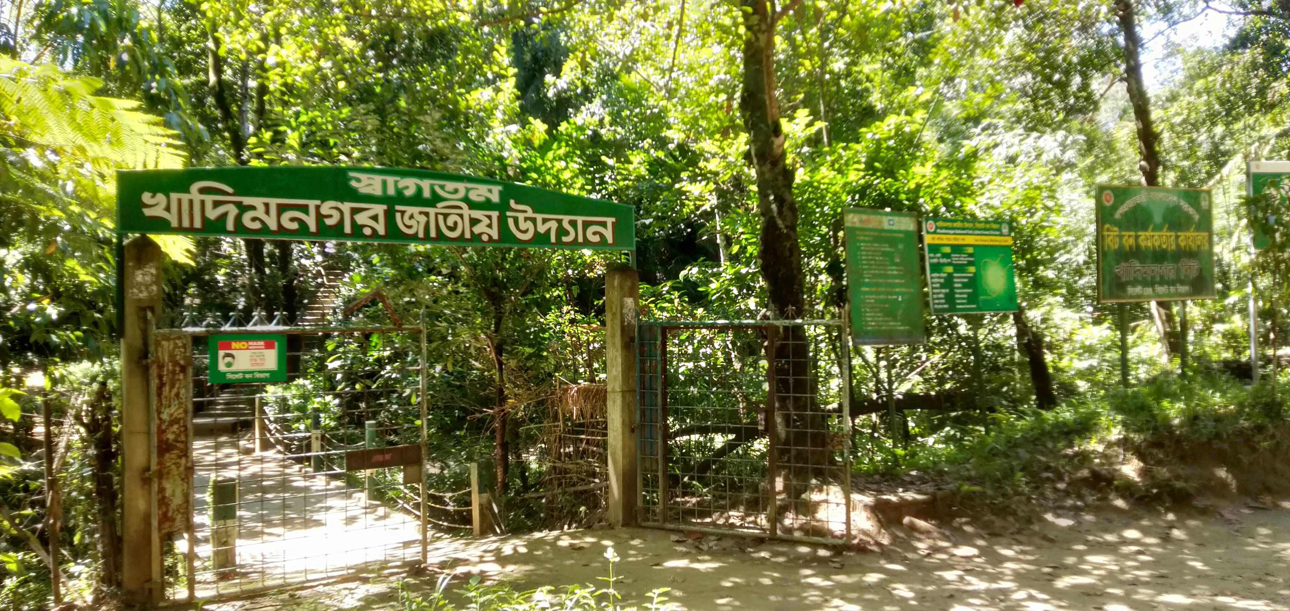 Green signboard with Bengali text marking the entrance to Khadim Nagar National Park, surrounded by trees and a dirt path