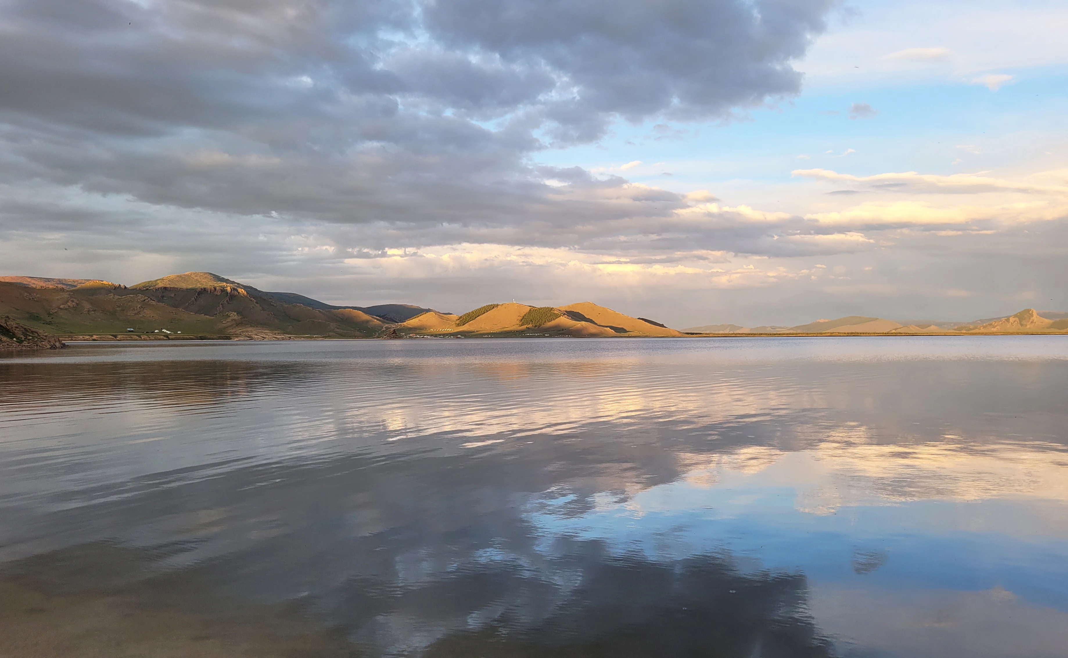 A wide, tranquil lake reflects a cloudy sunset sky, with gently rolling hills in the background.