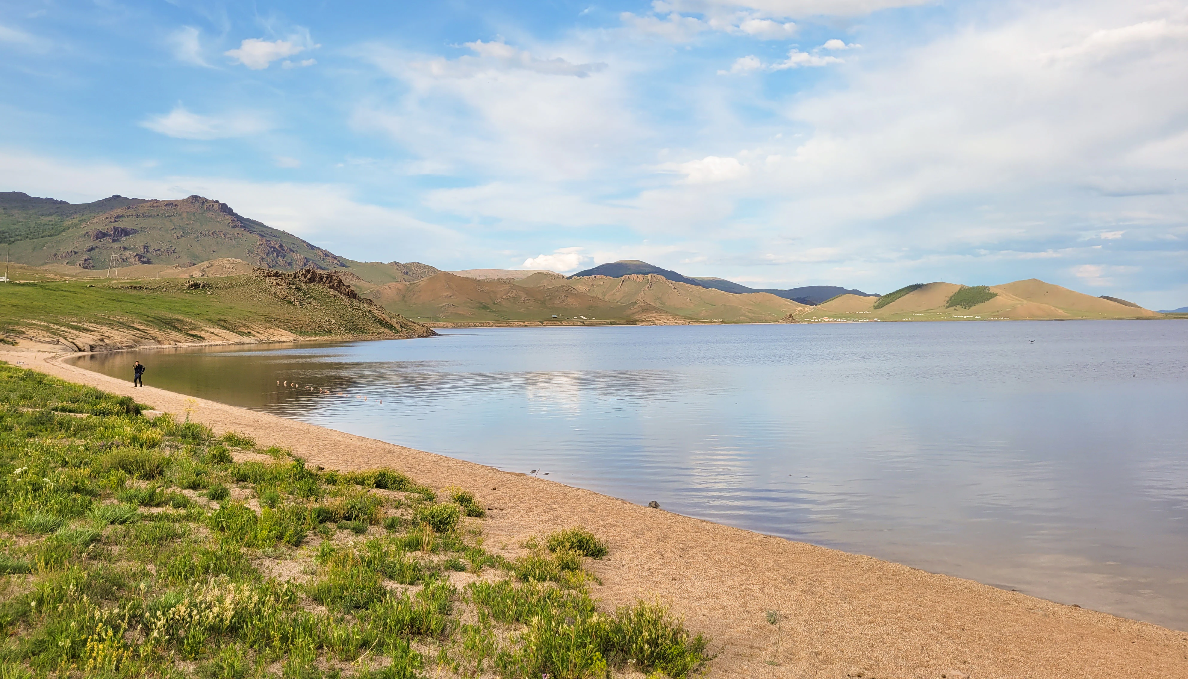 A wide view of a calm, large lake with a sandy shoreline, rolling hills in the background under a blue, partly cloudy sky.
