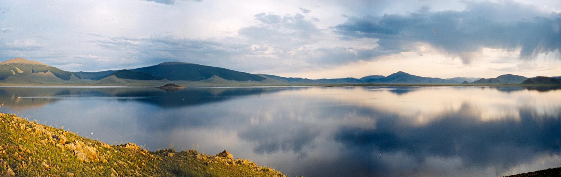 Panoramic landscape of a large, still lake reflecting the sky and clouds, framed by rolling green and rocky hills under a dramatic, partly cloudy sky.