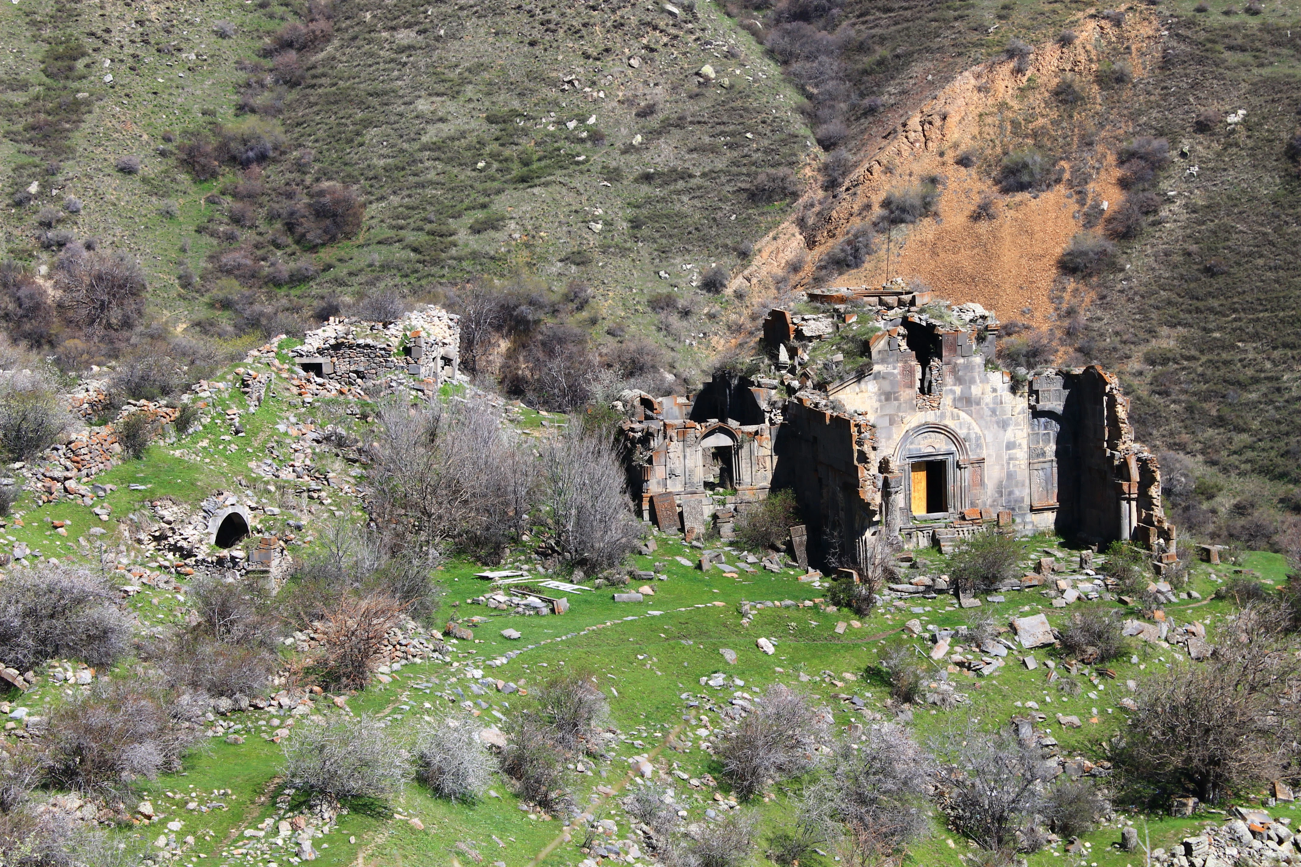 A high-angle view of the weathered stone ruins of Stepanos Monastery nestled in a green, rocky valley with steep, arid slopes in the background.