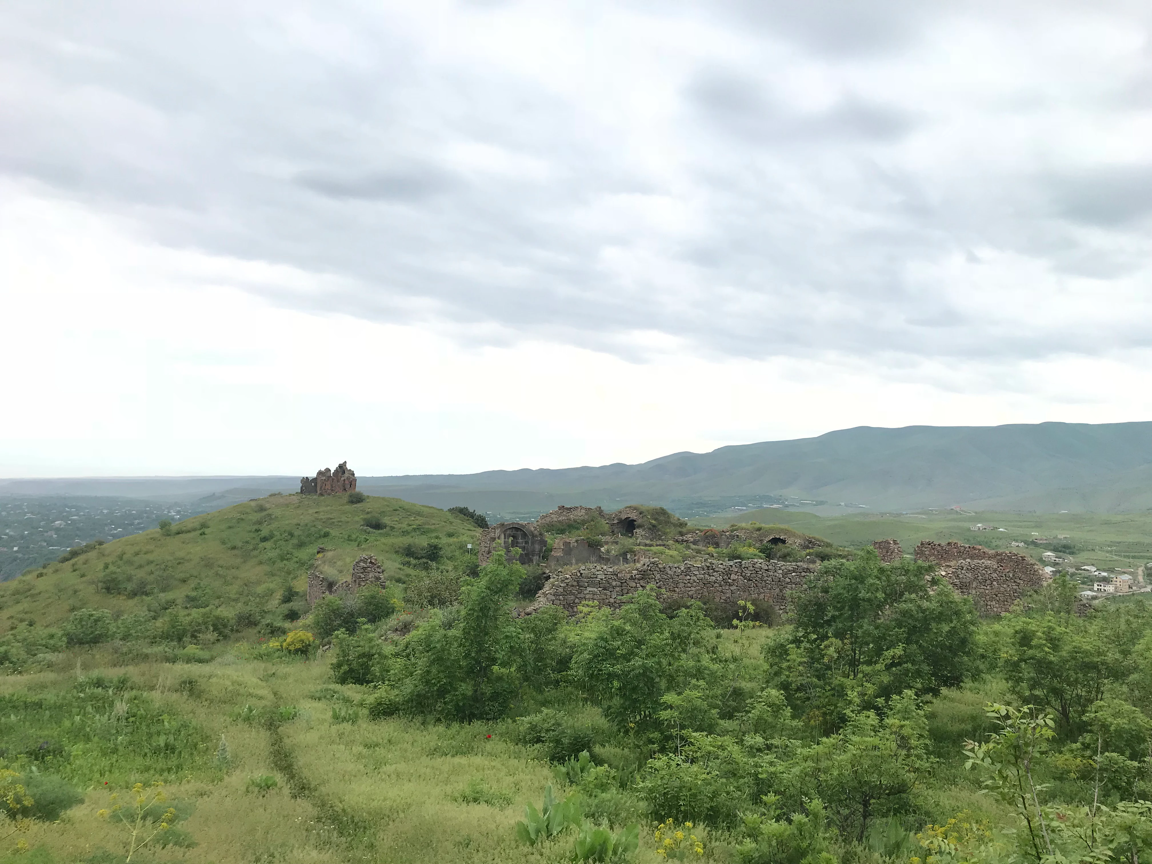A wide view of grassy, green hills under a cloudy sky featuring the ancient stone ruins of the Havuts Tar monastery.