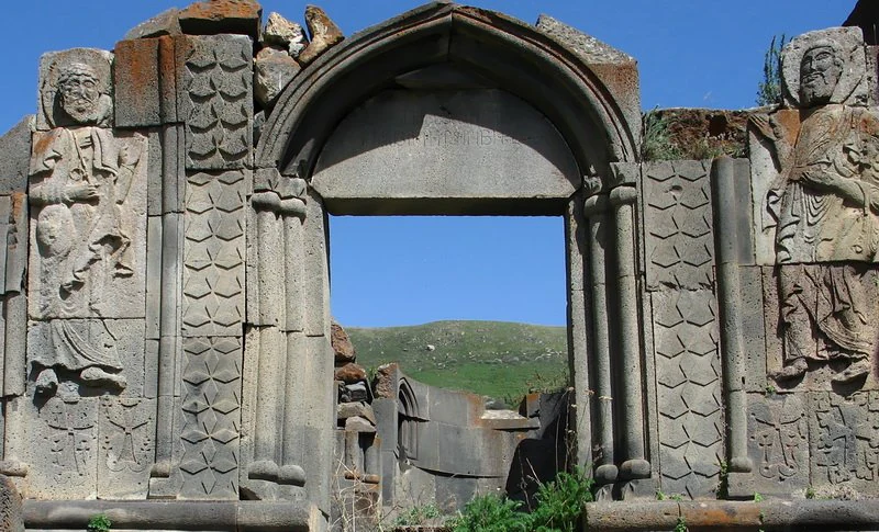Ancient stone doorway featuring detailed carved figures of saints and decorative masonry, framed against a clear blue sky and a distant grassy hill.
