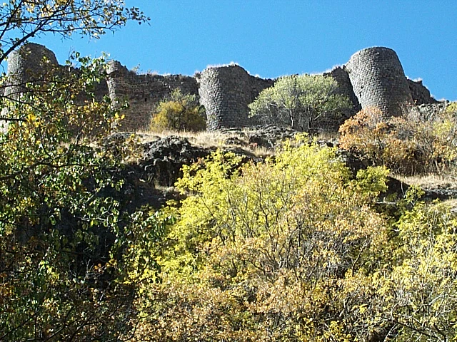 Ancient stone fortress ruins with prominent circular towers overlooking a vegetated hillside under a clear blue sky.