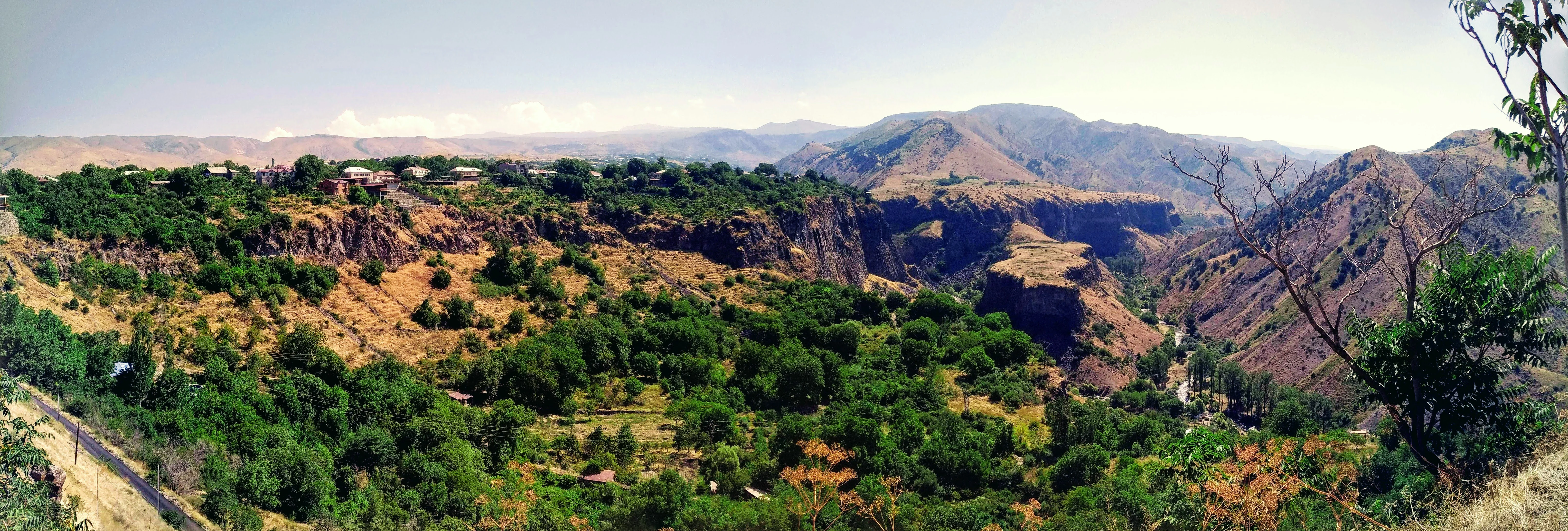 A panoramic landscape view showing a deep canyon with rocky cliffs and green forest canopy at the base, with arid hills in the background.