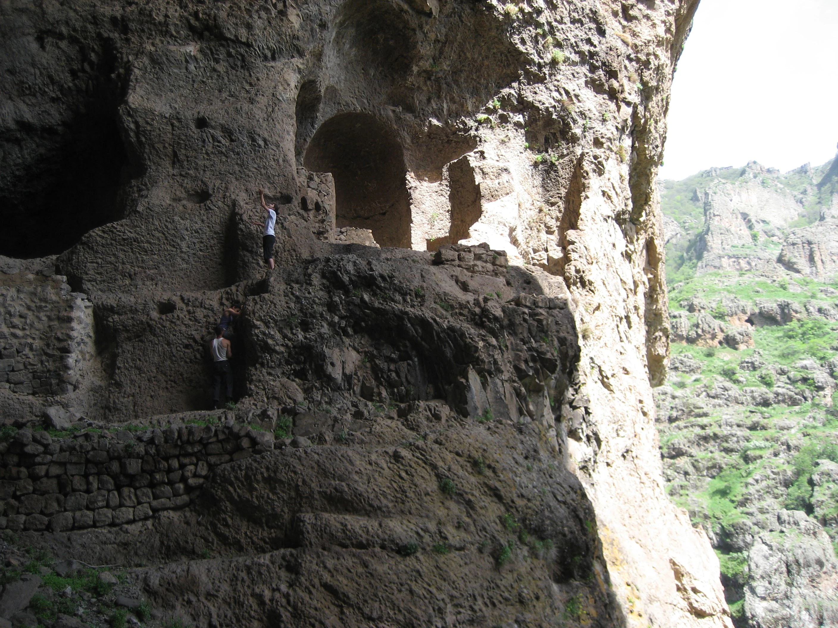 A view of ancient cave dwellings built directly into a steep, rugged rock face, with a canyon landscape visible in the background.
