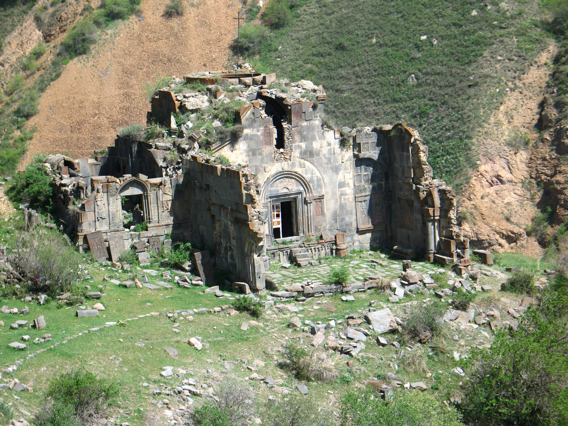 A high angle view of the ancient, ruined stone walls of the Aghjots Vank monastery, situated on a grassy slope with a rocky hillside in the background.