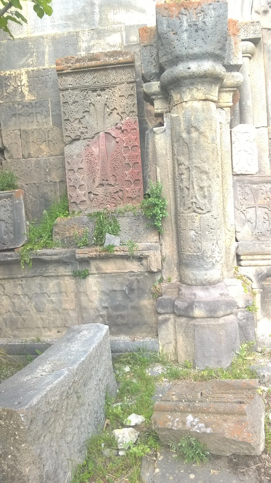 A weathered stone cross-stone, known as a khachkar, featuring intricate carvings, standing next to a carved stone pillar amidst ancient masonry ruins in a park setting.