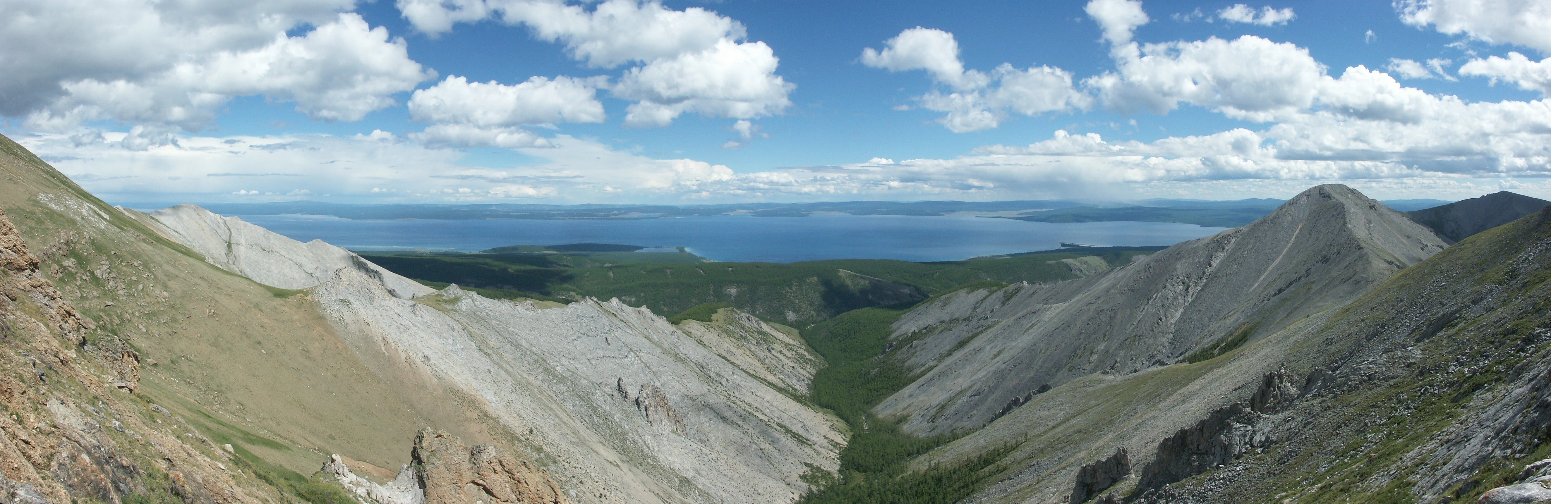 Panoramic landscape view from a high mountain ridge looking down into a forested valley towards a large, expansive blue lake under a sky with scattered white clouds.