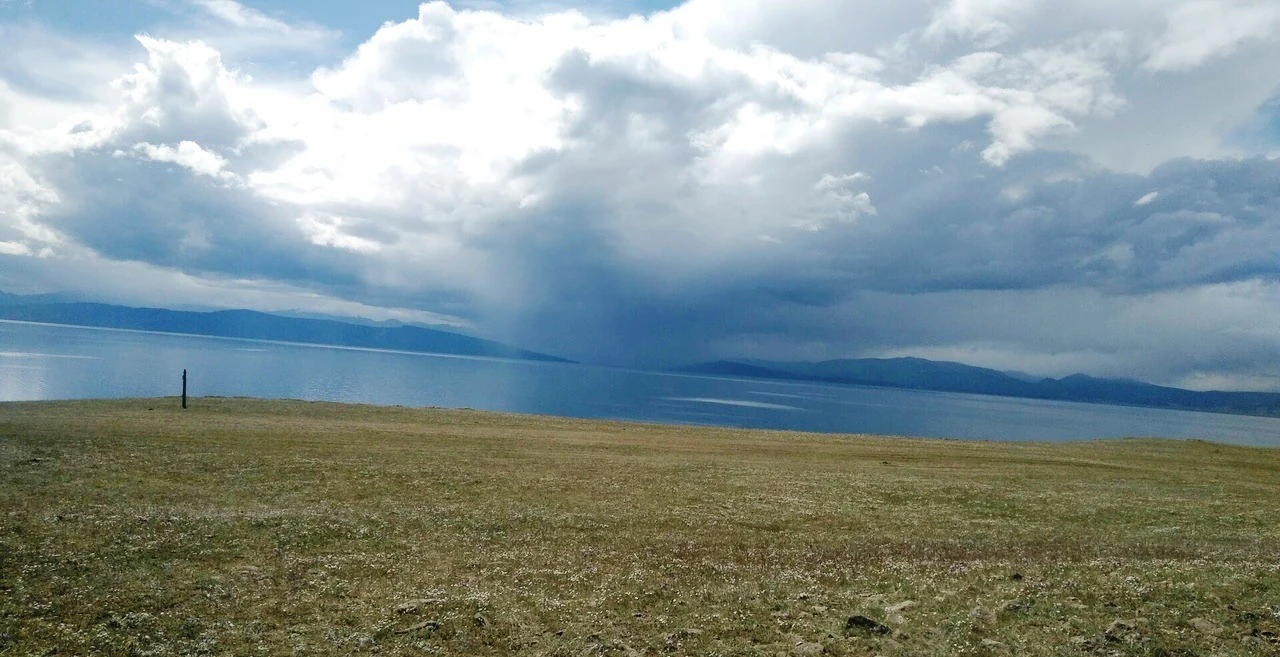 A wide view of a large blue lake under a sky filled with heavy, dark rain clouds; the foreground is a grassy, sloping field.