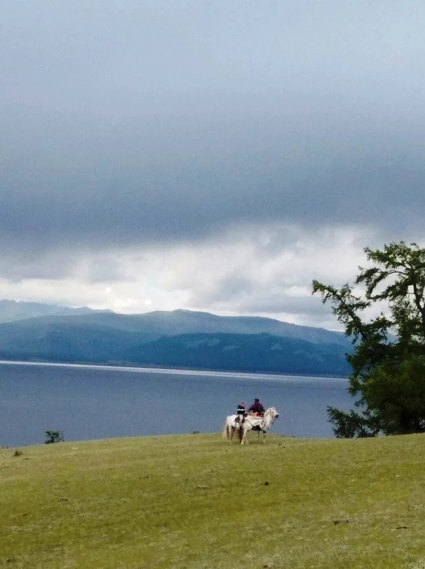 A wide view of a grassy hillside overlooking a large lake and distant mountains under a cloudy sky, with a person riding a white horse in the foreground.
