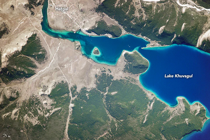 Satellite image showing the deep blue water of Lake Khövsgöl, the village of Hatgal at the shoreline, and surrounding dense boreal forests and rocky terrain.