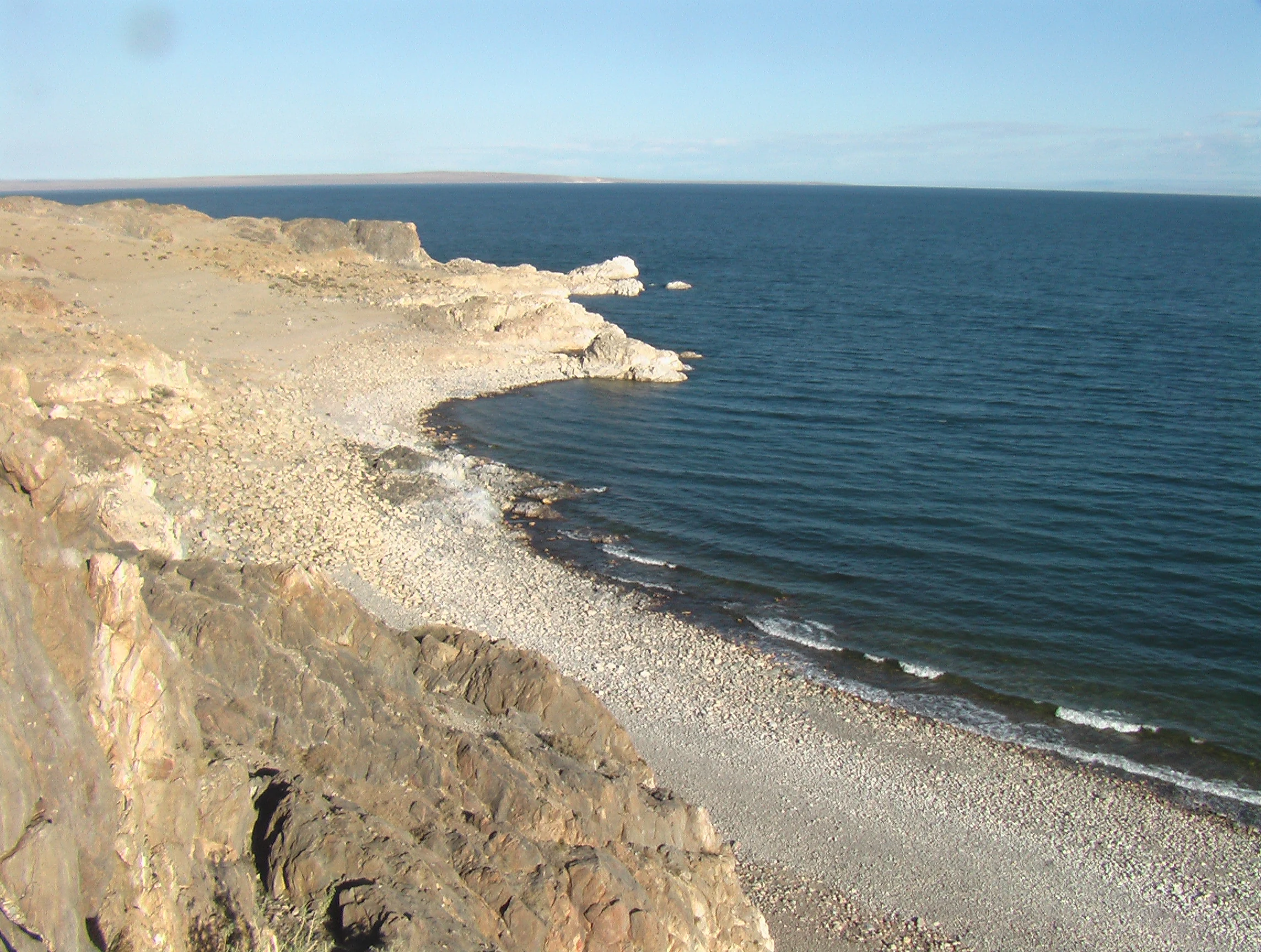 A wide view of the rocky, arid shoreline of Khyargas Lake in Mongolia, with deep blue water meeting a pebble-covered coast under a clear sky.