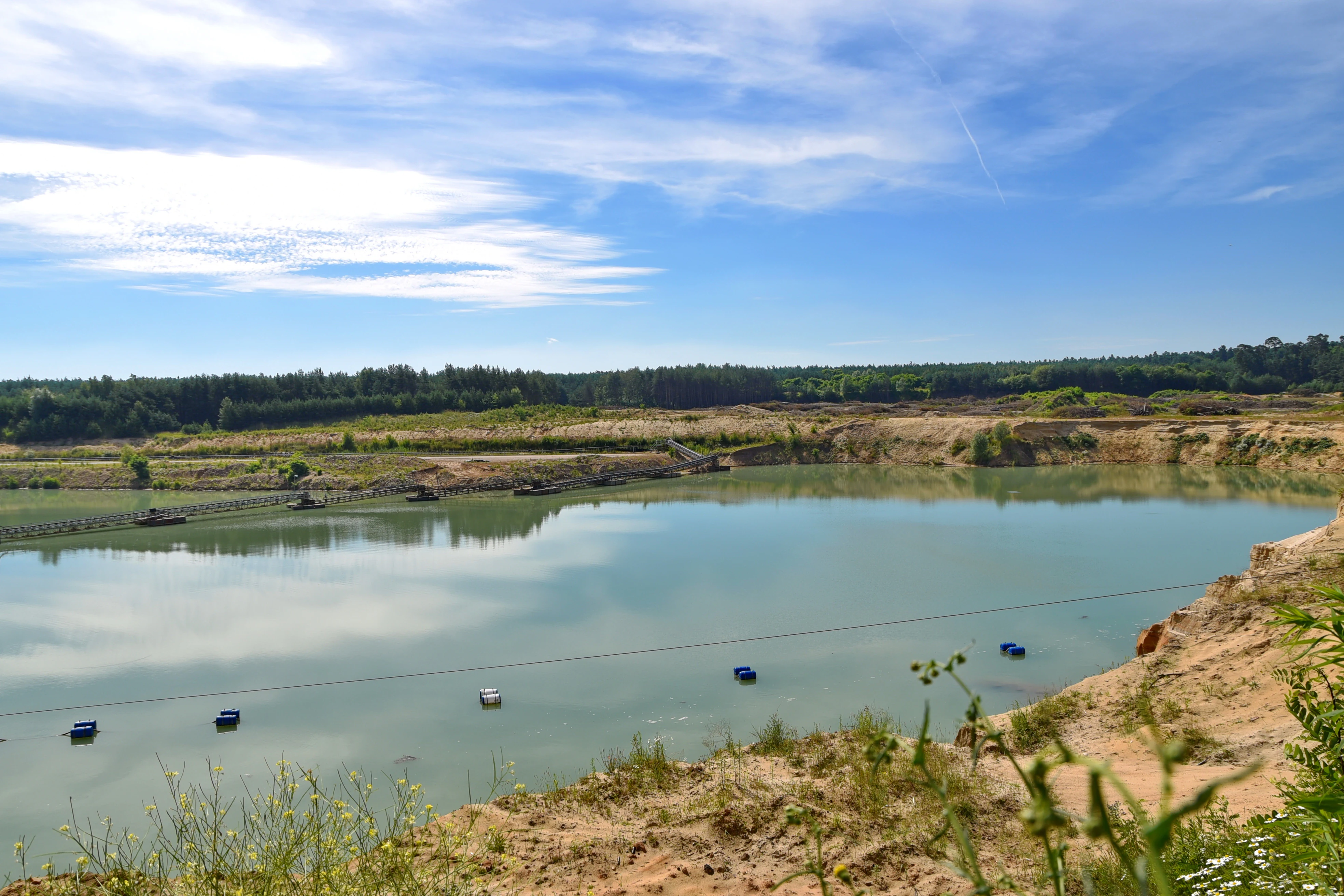 A wide view of a turquoise pond within an old gravel and sand pit, surrounded by sandy slopes and a forest line under a blue sky.