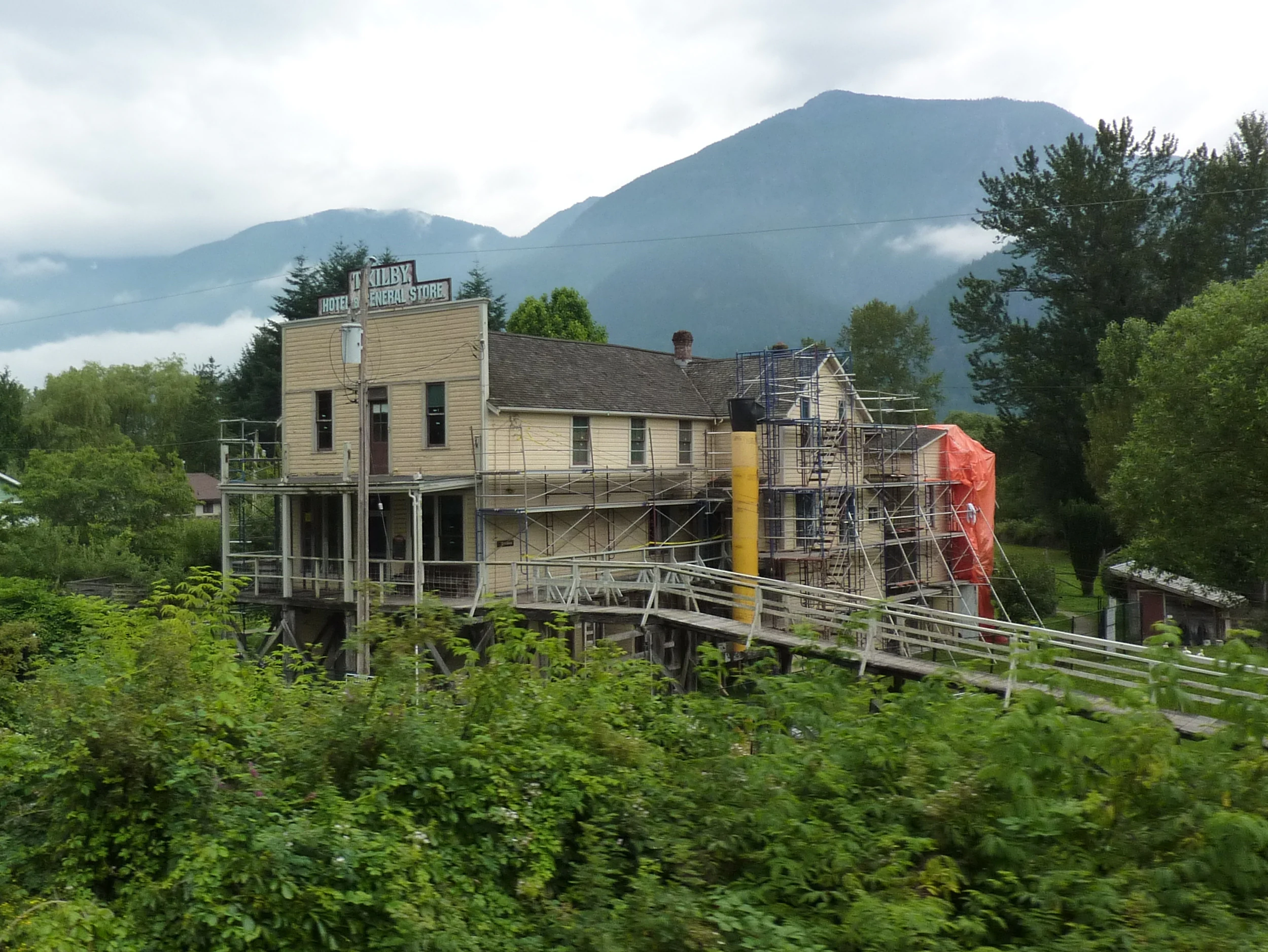 A historic, two-story wooden building with scaffolding surrounding it, set against a backdrop of hazy, cloud-covered mountains.