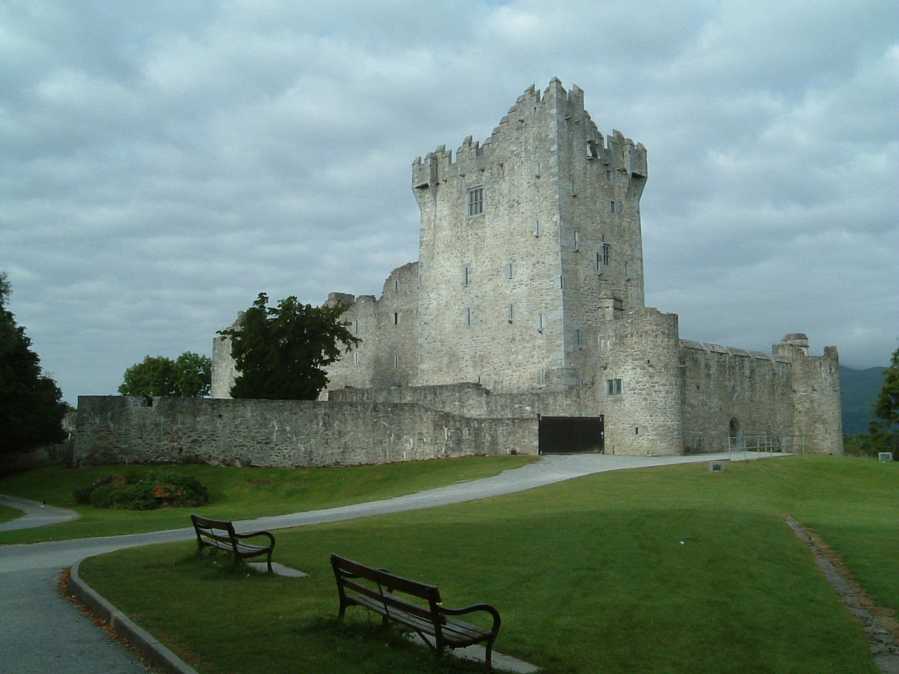 Stone castle with multiple towers and surrounding walls in a grassy area with paved pathways and benches