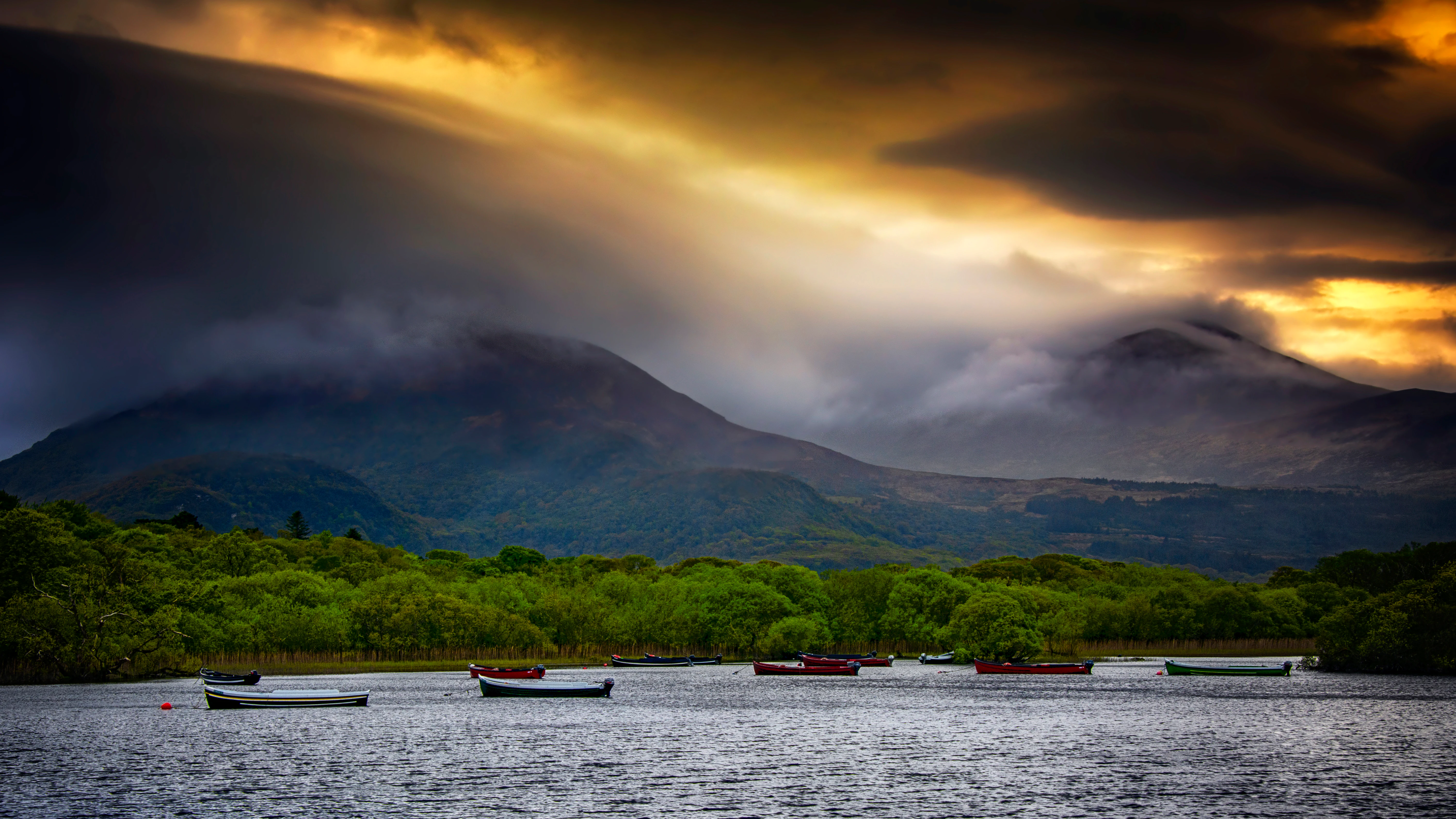 A lake with several small boats on calm water, surrounded by dense green forests and mountains under a cloudy sky with sunlight breaking through