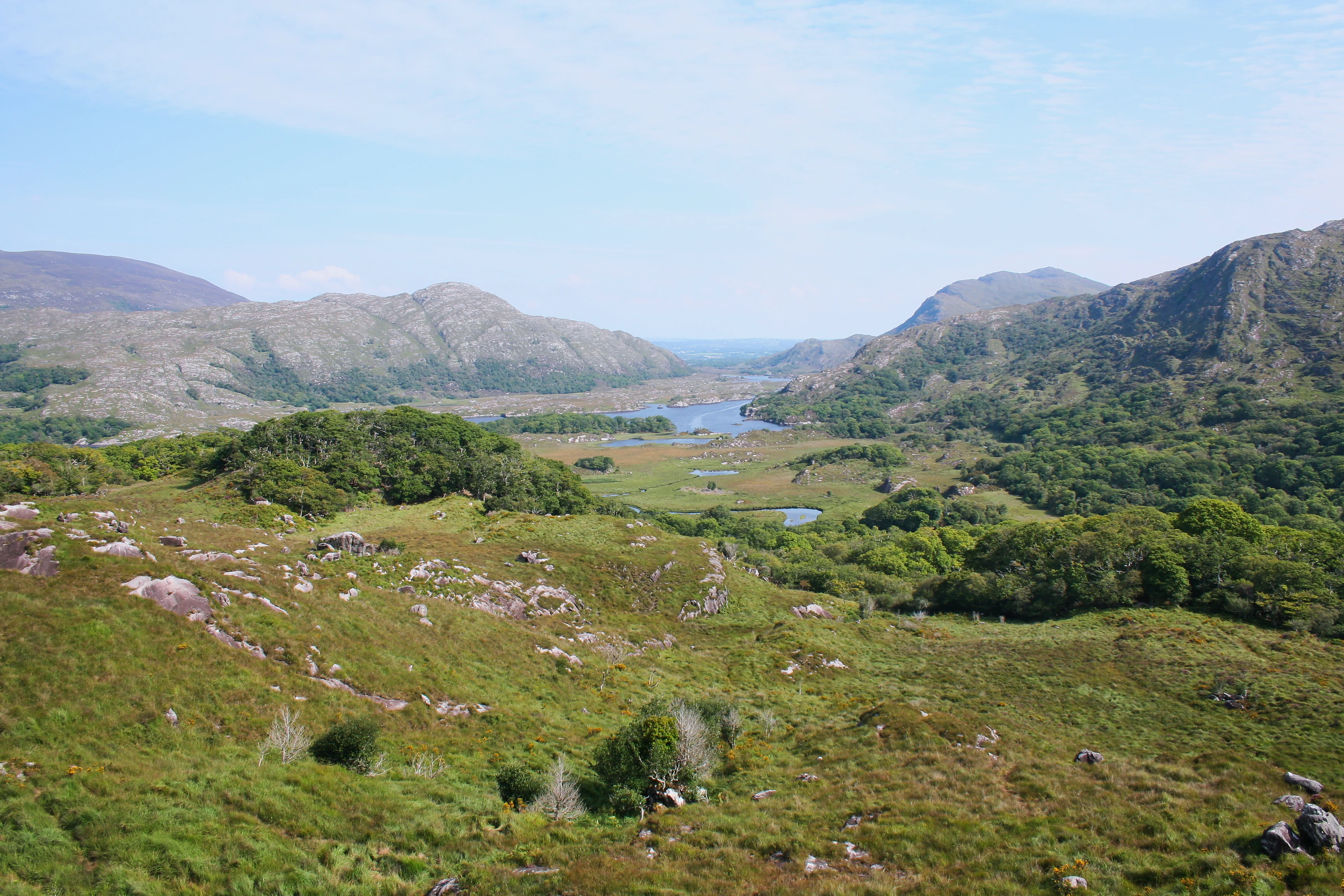 Landscape view of rolling green hills, rocky outcrops, forests, and lakes in the distance under a partly cloudy sky