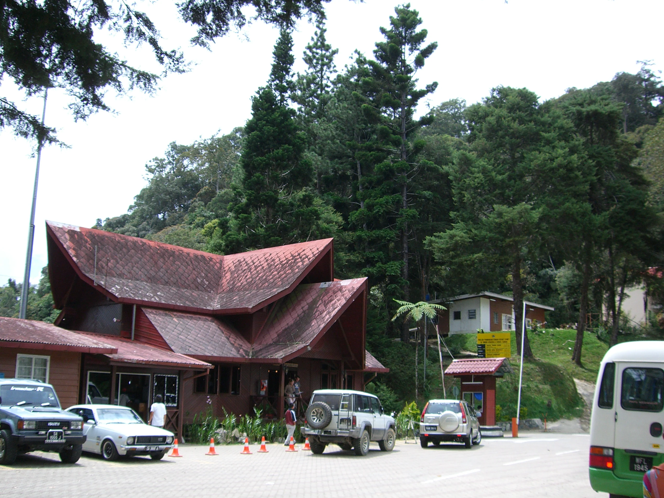 A view of the Kinabalu Park headquarters building, characterized by its traditional-style red roof, with several parked vehicles in the foreground and tall green trees in the background.