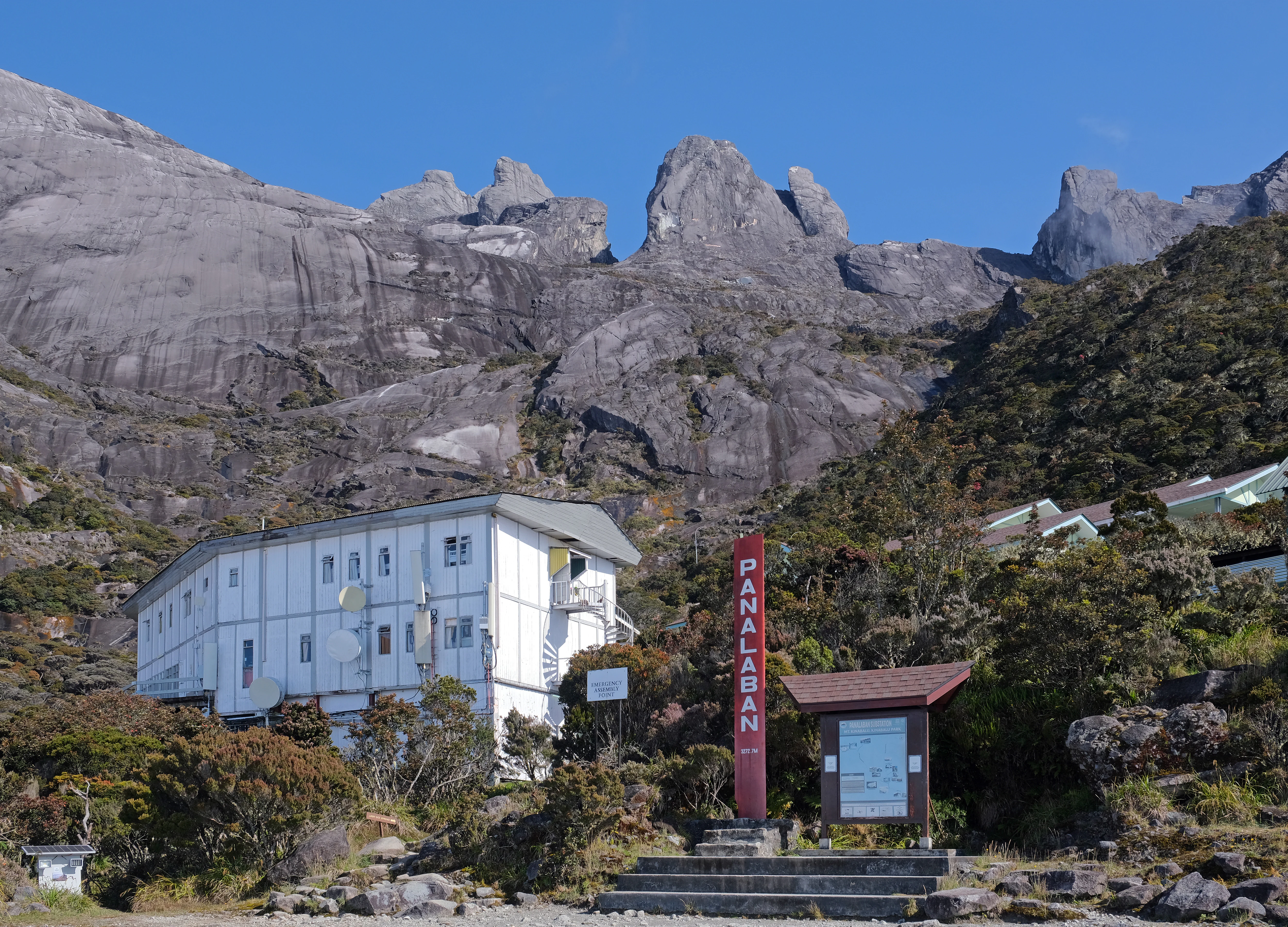 A white multi-story building sits at the base of steep rocky mountain peaks with a red sign labeled PANALABAN nearby.