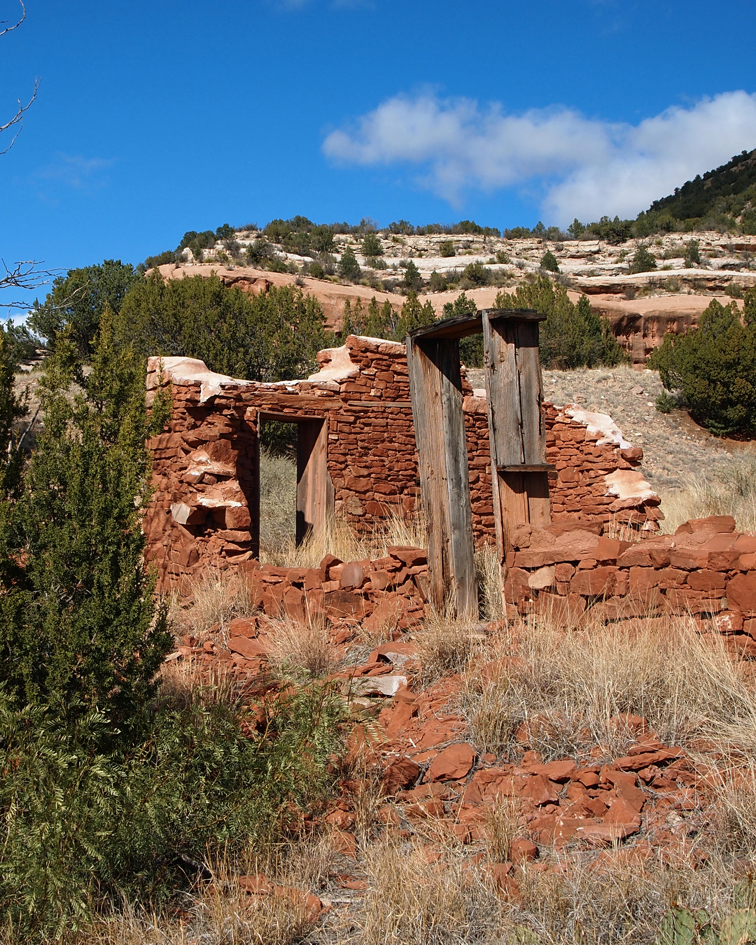 A vertical view of stone and wood building ruins in a desert canyon landscape under a blue sky.